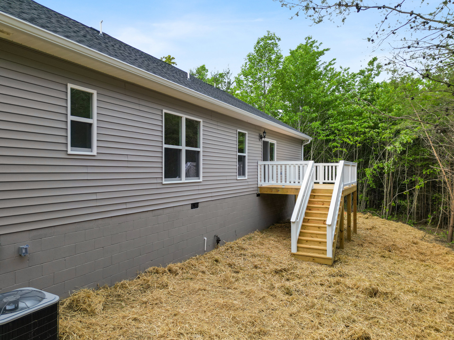 Two-story home with gray siding, elevated wooden deck, white-framed windows, exterior staircase, rooftop partially visible, air conditioning unit, surrounded by grass and mature