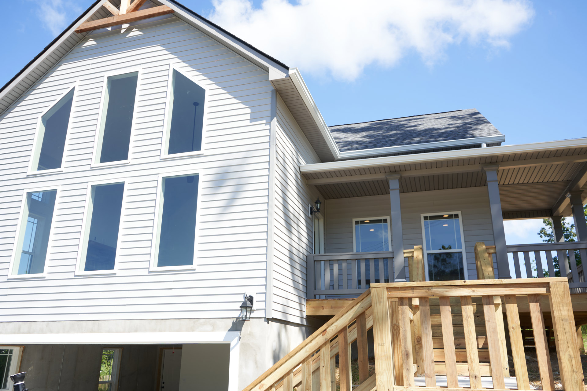 Two-story home exterior with wood siding, prominent wooden staircase and railing, illuminated window, gray roof, and blue sky with scattered clouds