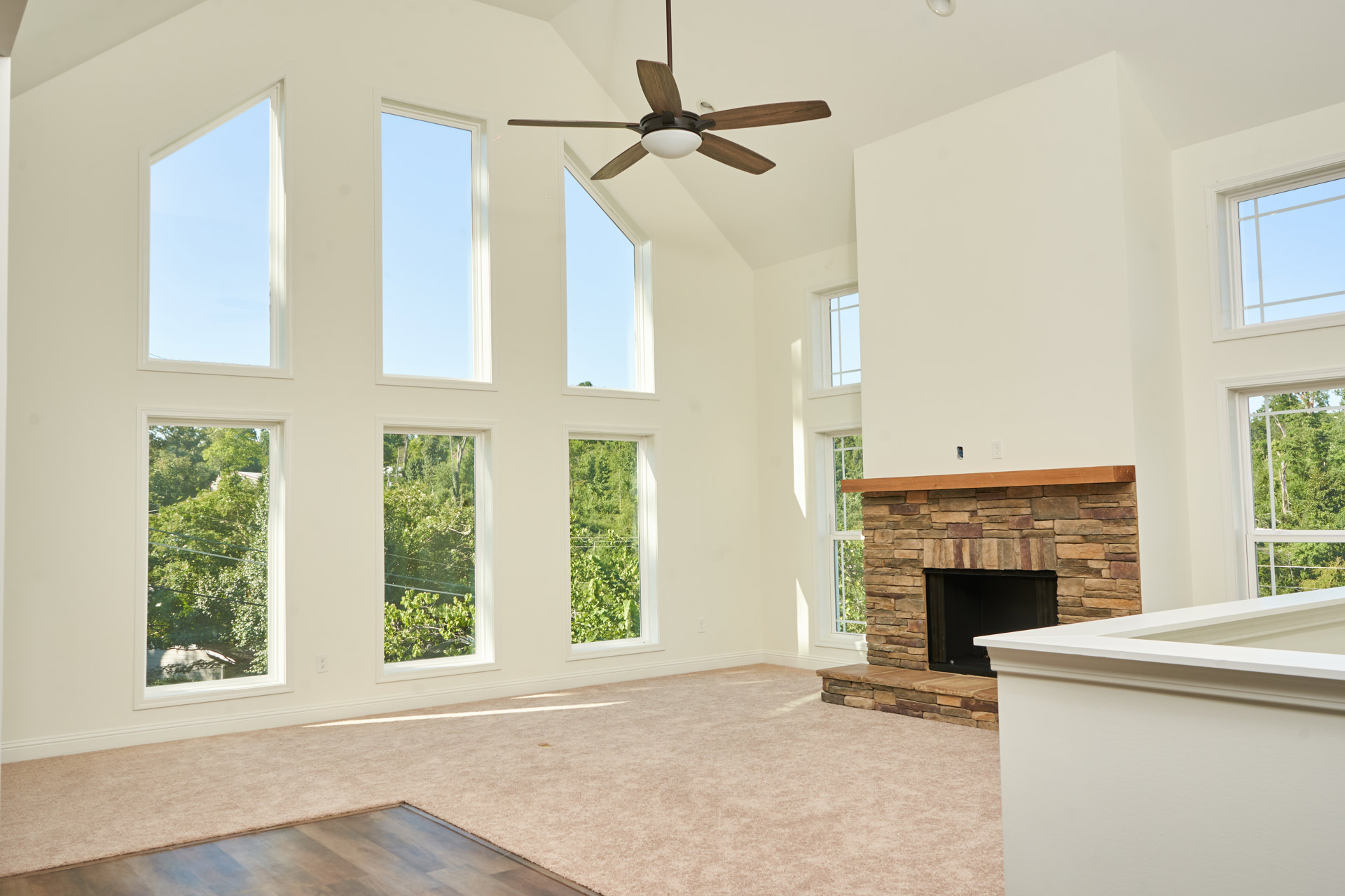 Living room featuring a white fireplace with stone surround, ceiling fan with light fixture, large windows letting in natural light, neutral walls, and hardwood flooring