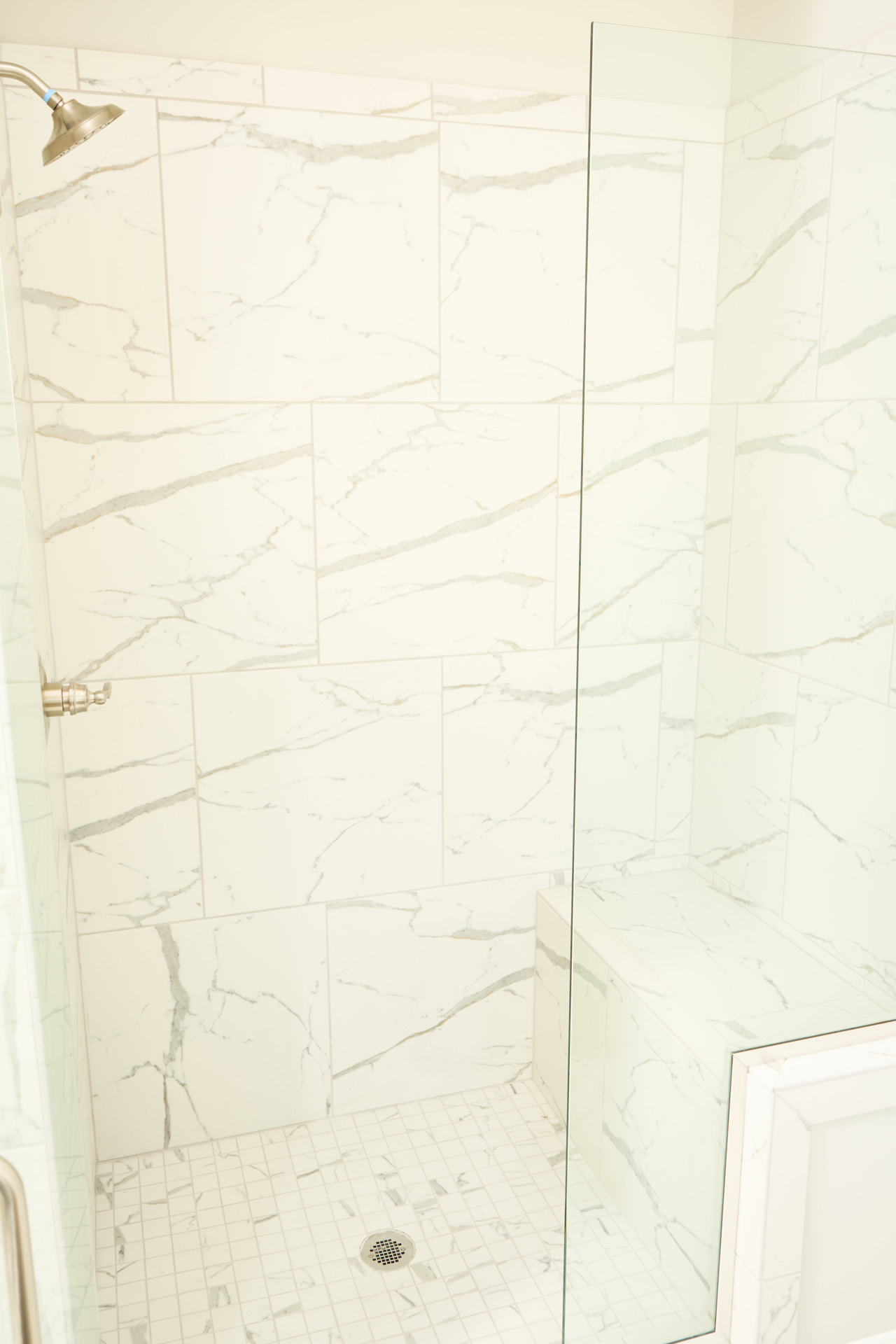 White tile shower with glass door, chrome shower head featuring a blue accent, metal drain cover, and white tiled wall