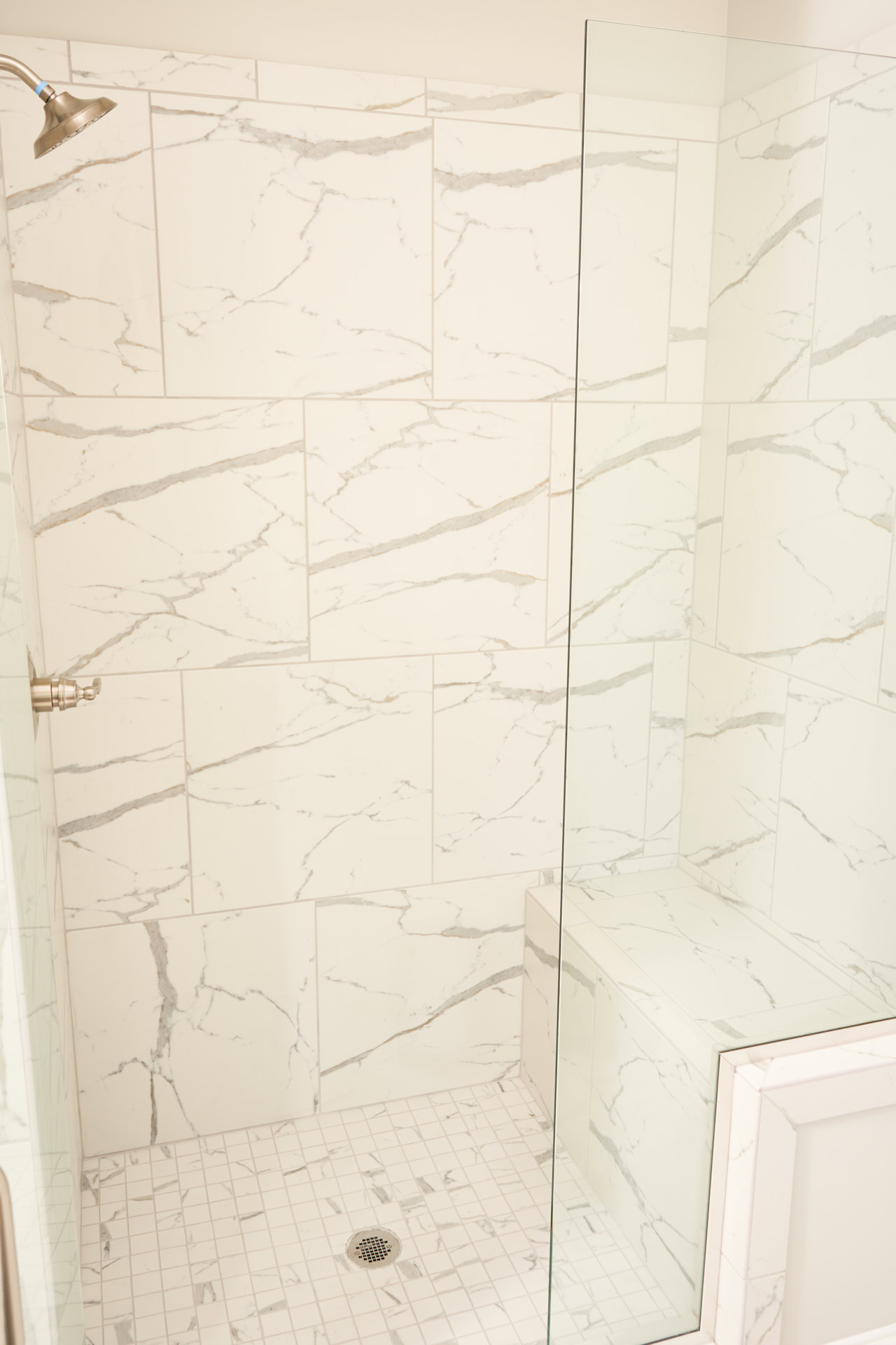 White tiled shower with silver framed glass door, chrome shower head featuring a blue accent, close-up of round drain, white corner wall, detailed finial, and white tile with