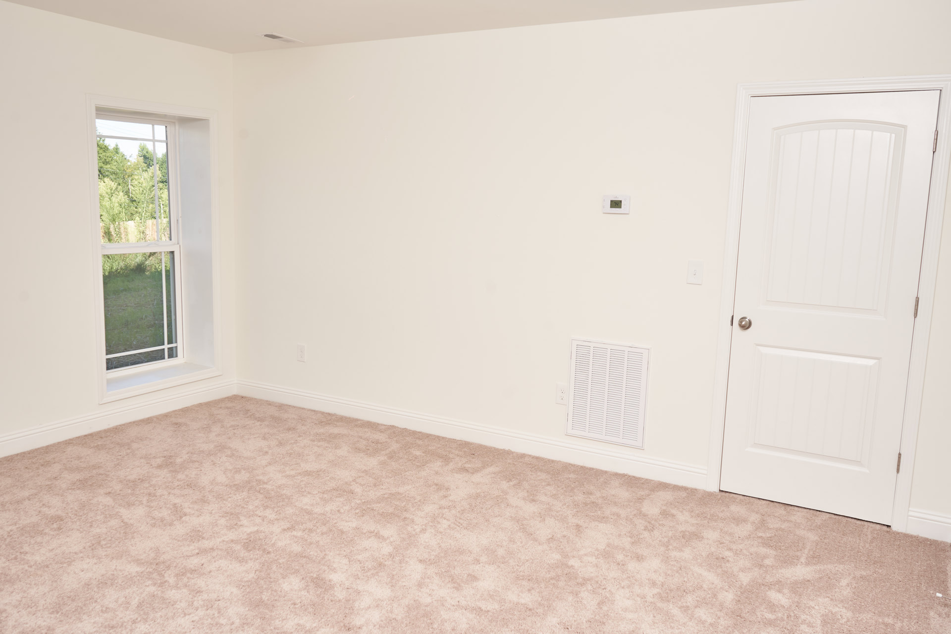 Carpeted bedroom with white walls, white door featuring a silver handle, rectangular window overlooking trees and grass, and a white wall vent