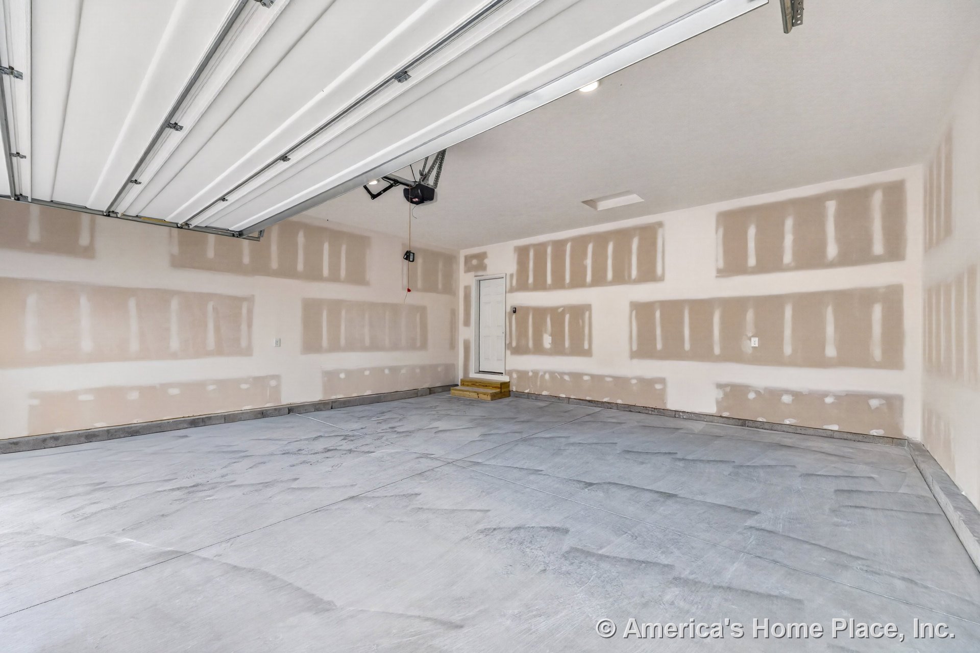 Unfinished drywall walls and concrete slab flooring in a residential garage, overhead garage door with ceiling-mounted opener, recessed ceiling light, white entry door accessed by