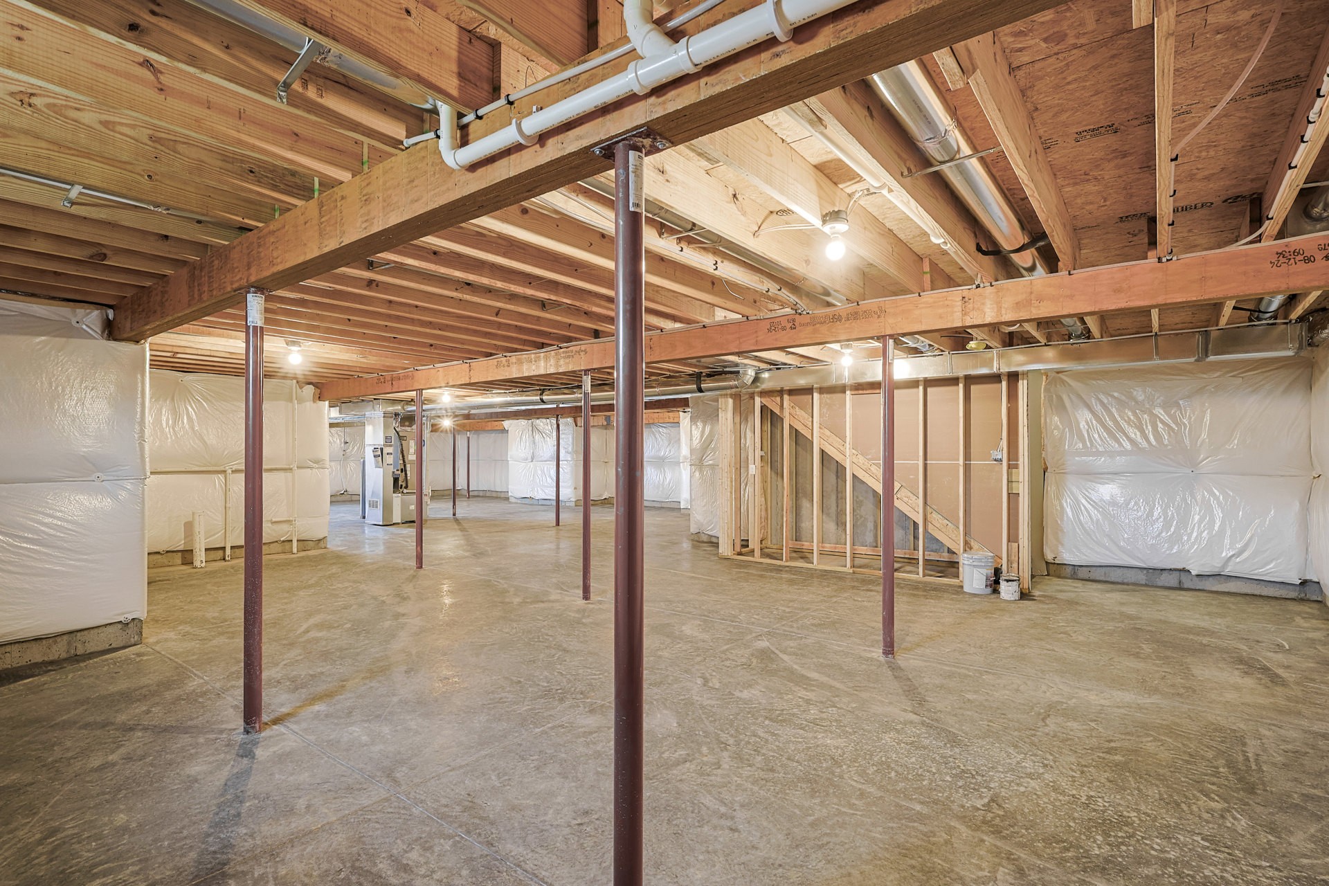 Basement with exposed wooden ceiling beams, metal support pole, visible pipes, white plastic wall covering, concrete floor