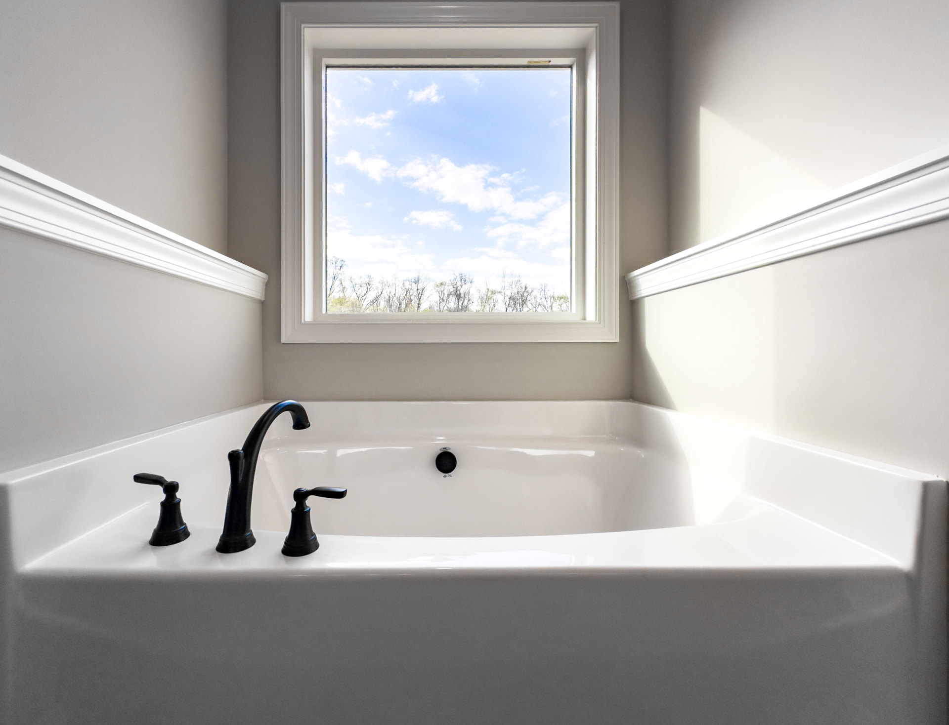 White freestanding bathtub beneath large window with sky and treetop view, matte black faucet and handle, light-colored walls, minimalist bathroom decor