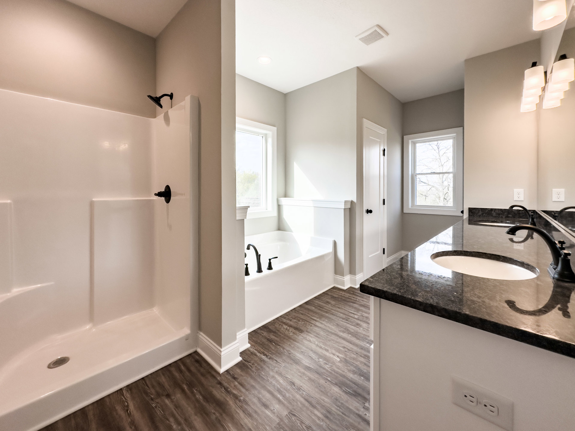 White freestanding bathtub with matte black shower head, white vessel sink on vanity, large window framing leafy tree, light tile walls and floor, black faucet fixtures