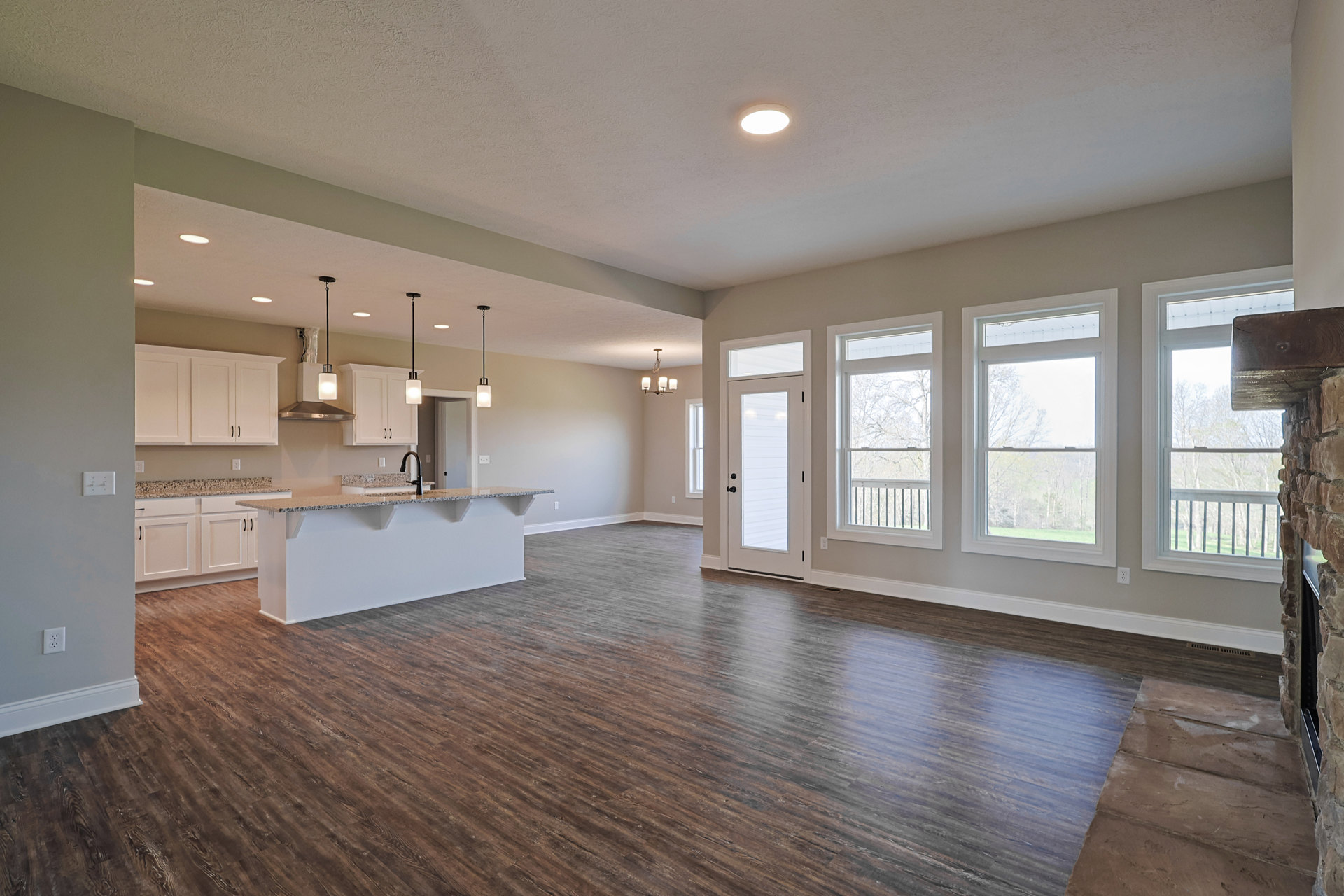 Spacious kitchen with white walls, hardwood flooring, central island, ceiling light fixture, large windows overlooking trees, and a door leading outside