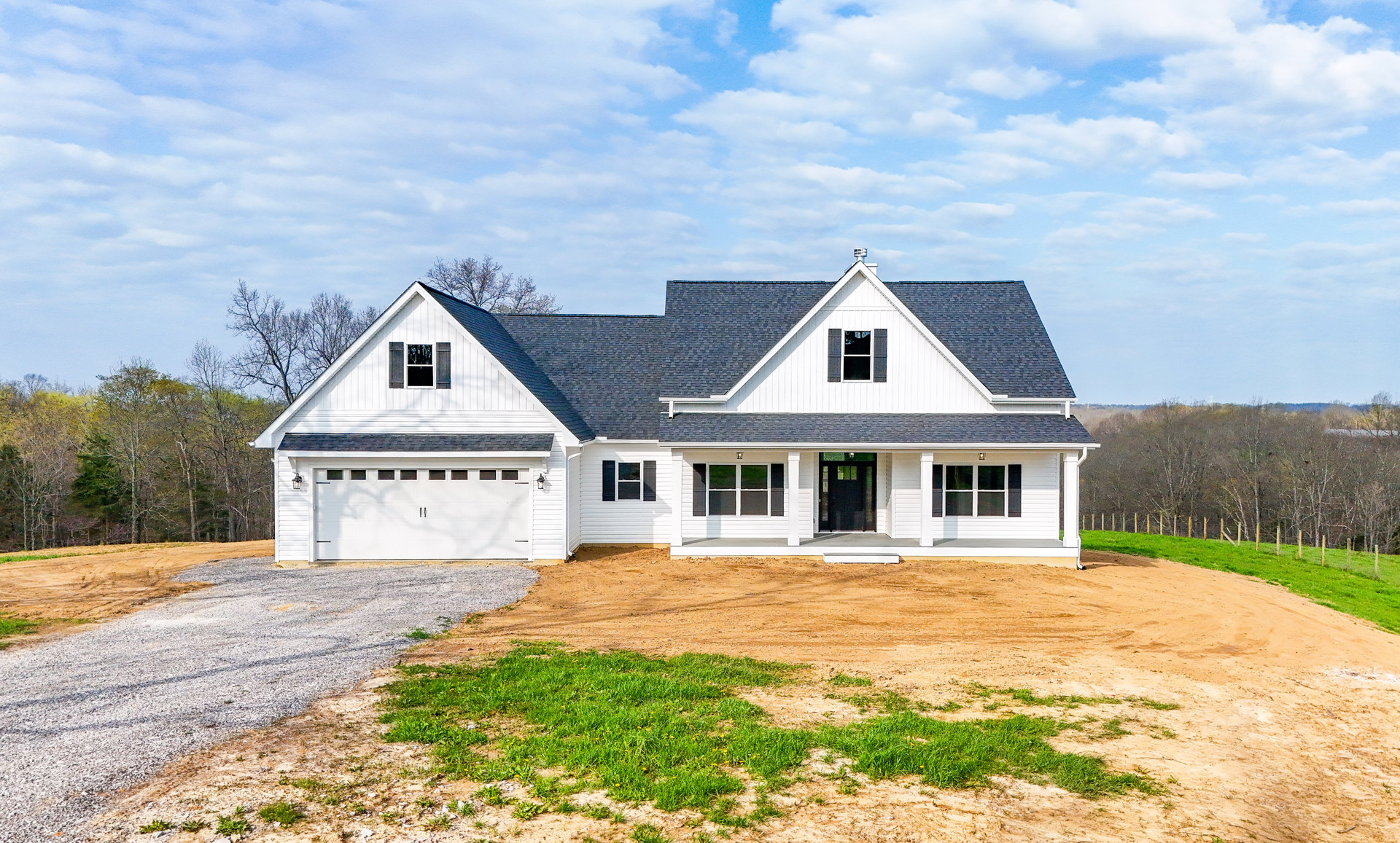 White house with black roof and black glass-paneled door, white-framed windows, concrete driveway, green grass lawn, blue sky with scattered clouds
