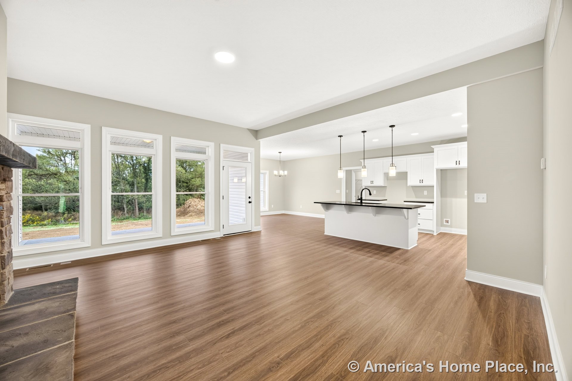 Four tall windows line the living room with wood-look plank flooring, open layout flows into kitchen featuring white shaker cabinets, black countertops, kitchen island with pendant