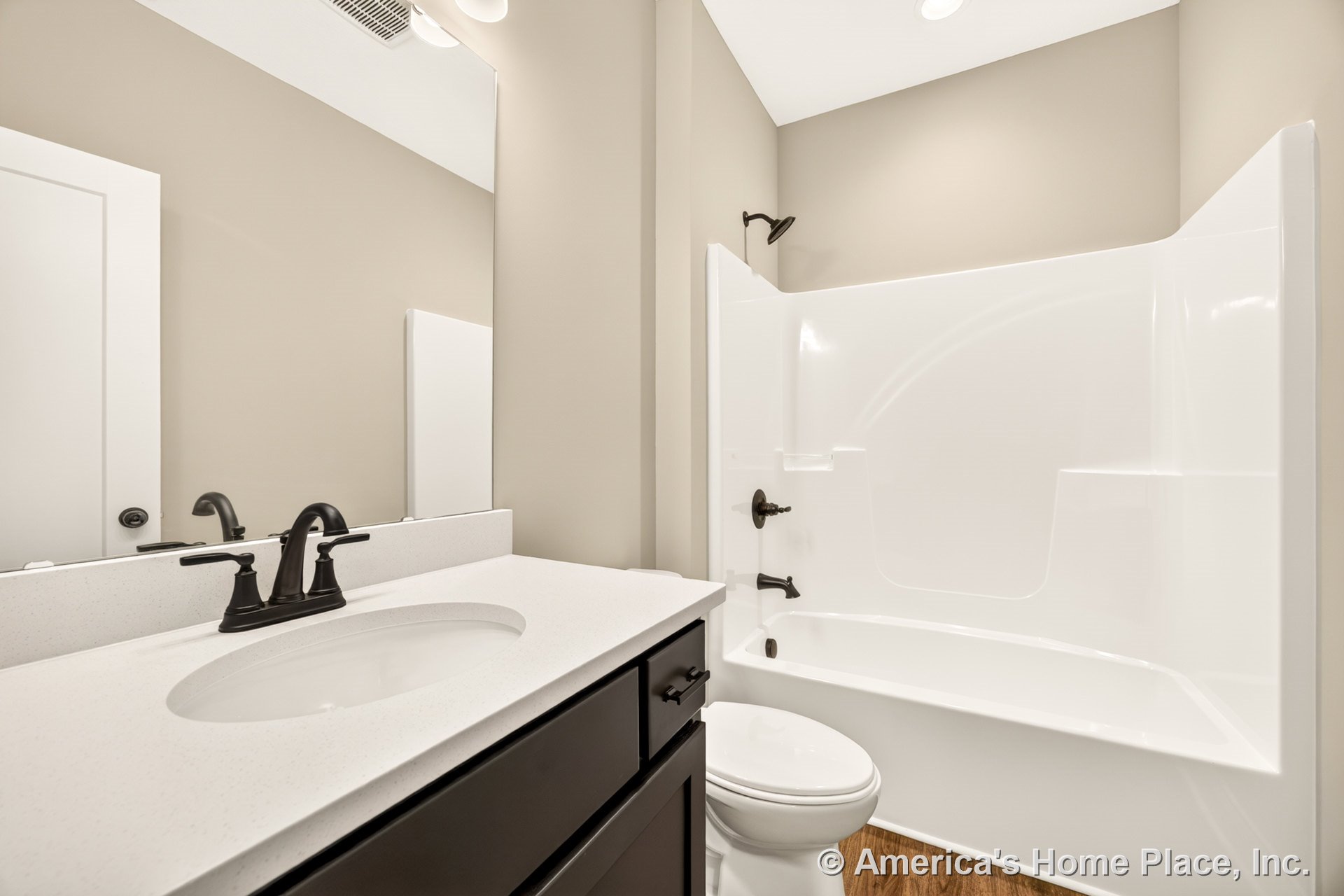 Built-in bathtub with integrated shower, dark vanity cabinet topped by white solid surface countertop, large wall mirror, matte black plumbing fixtures, wood-look flooring