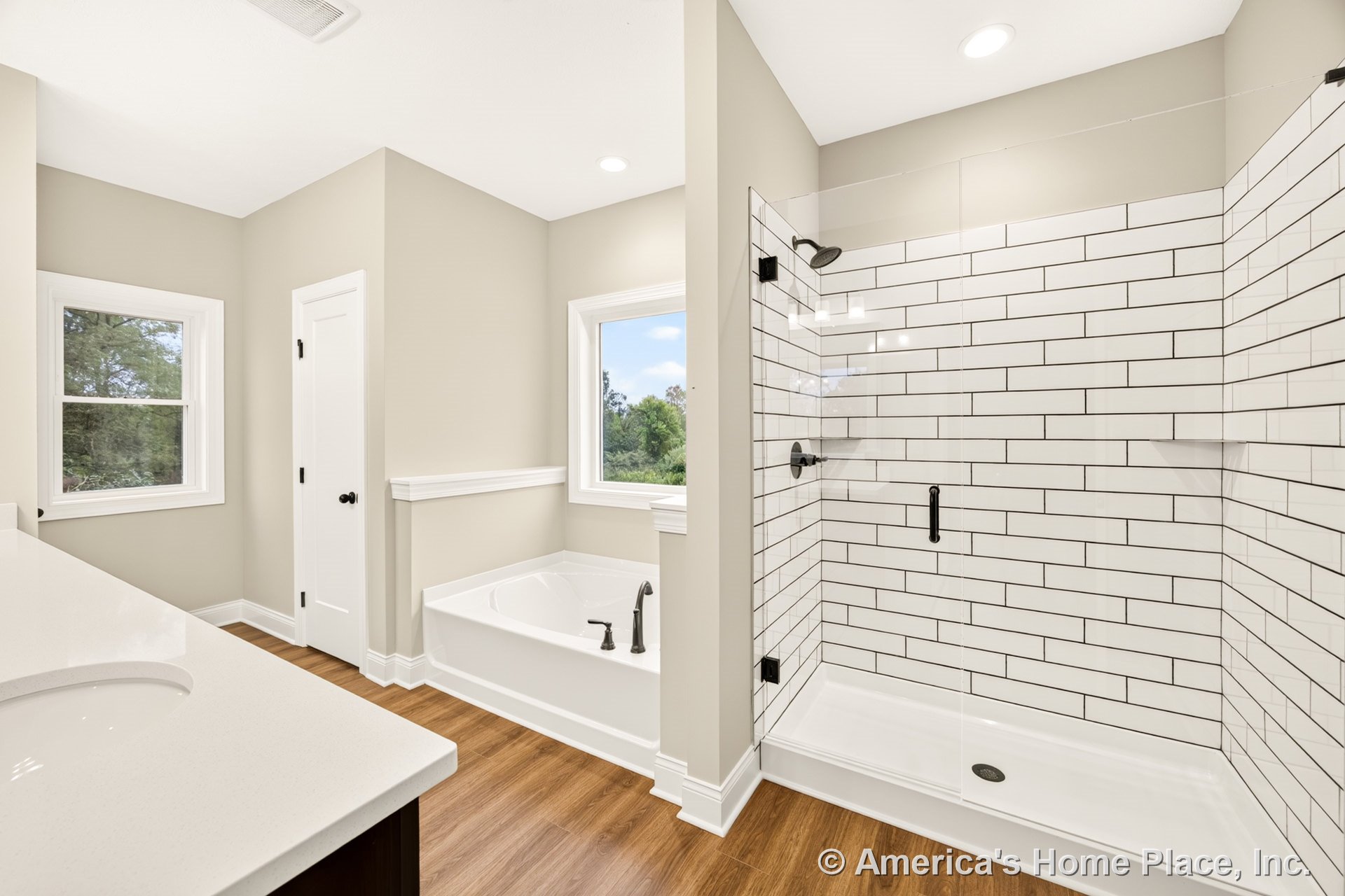 Walk-in shower with glass enclosure and white subway tile walls, built-in soaking tub, double vanity topped with quartz countertop, wood-look flooring, recessed ceiling lights