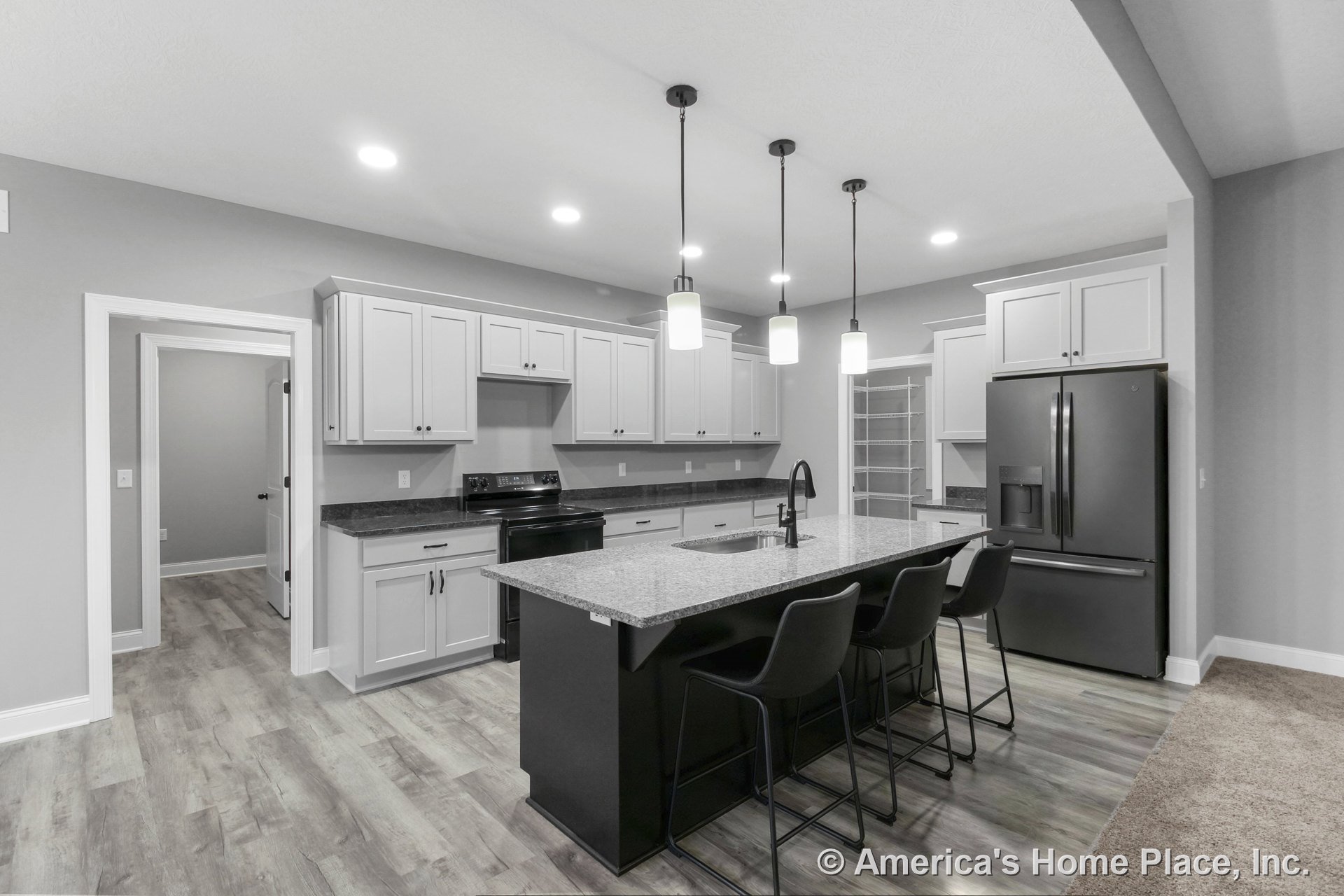 Spacious kitchen with large granite island countertop, white shaker cabinets, black refrigerator and appliances, pendant lighting above island, walk-in pantry with built-in
