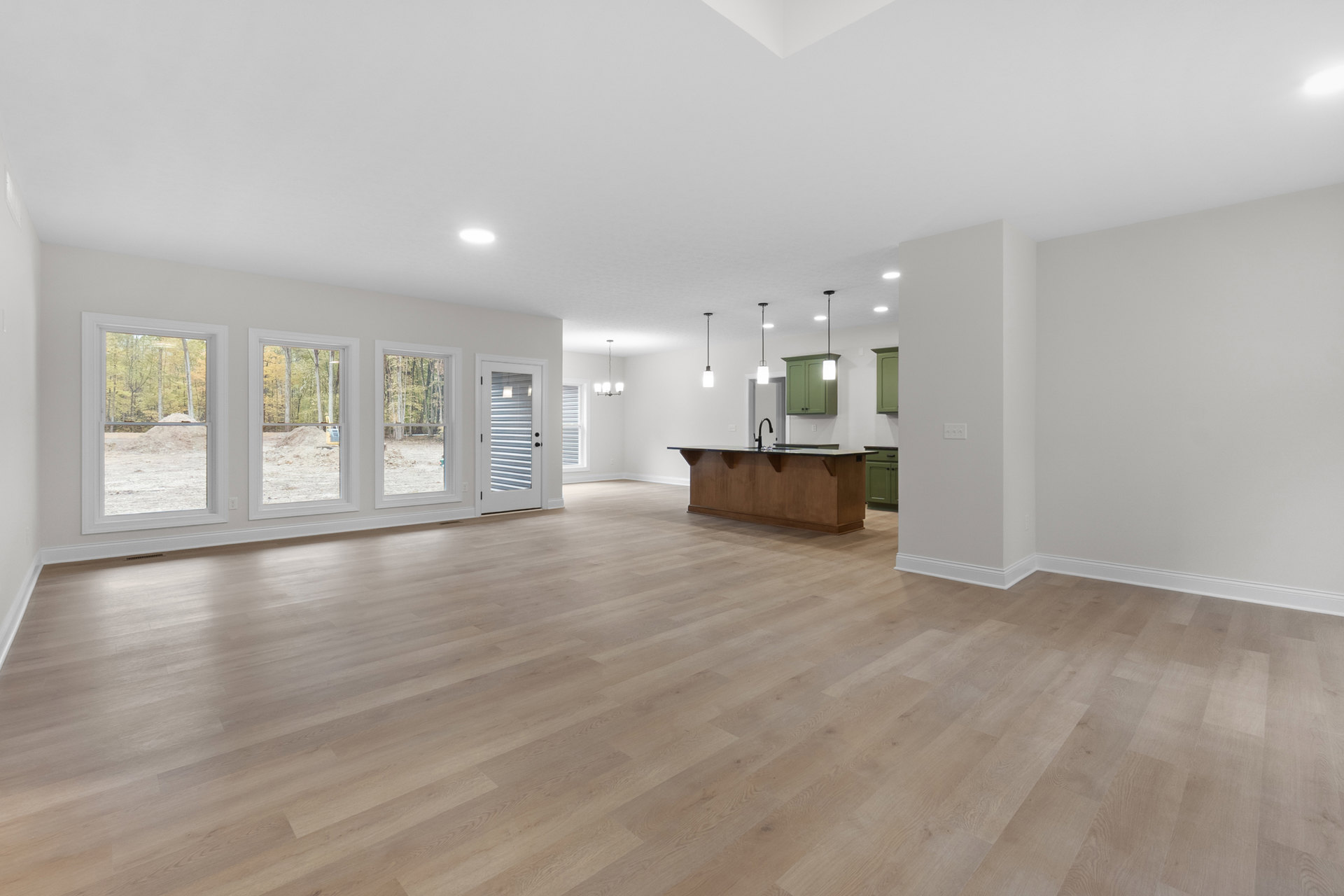 Open-plan room with hardwood flooring, modern kitchen featuring wood cabinetry and black countertop, white door, large window overlooking trees.