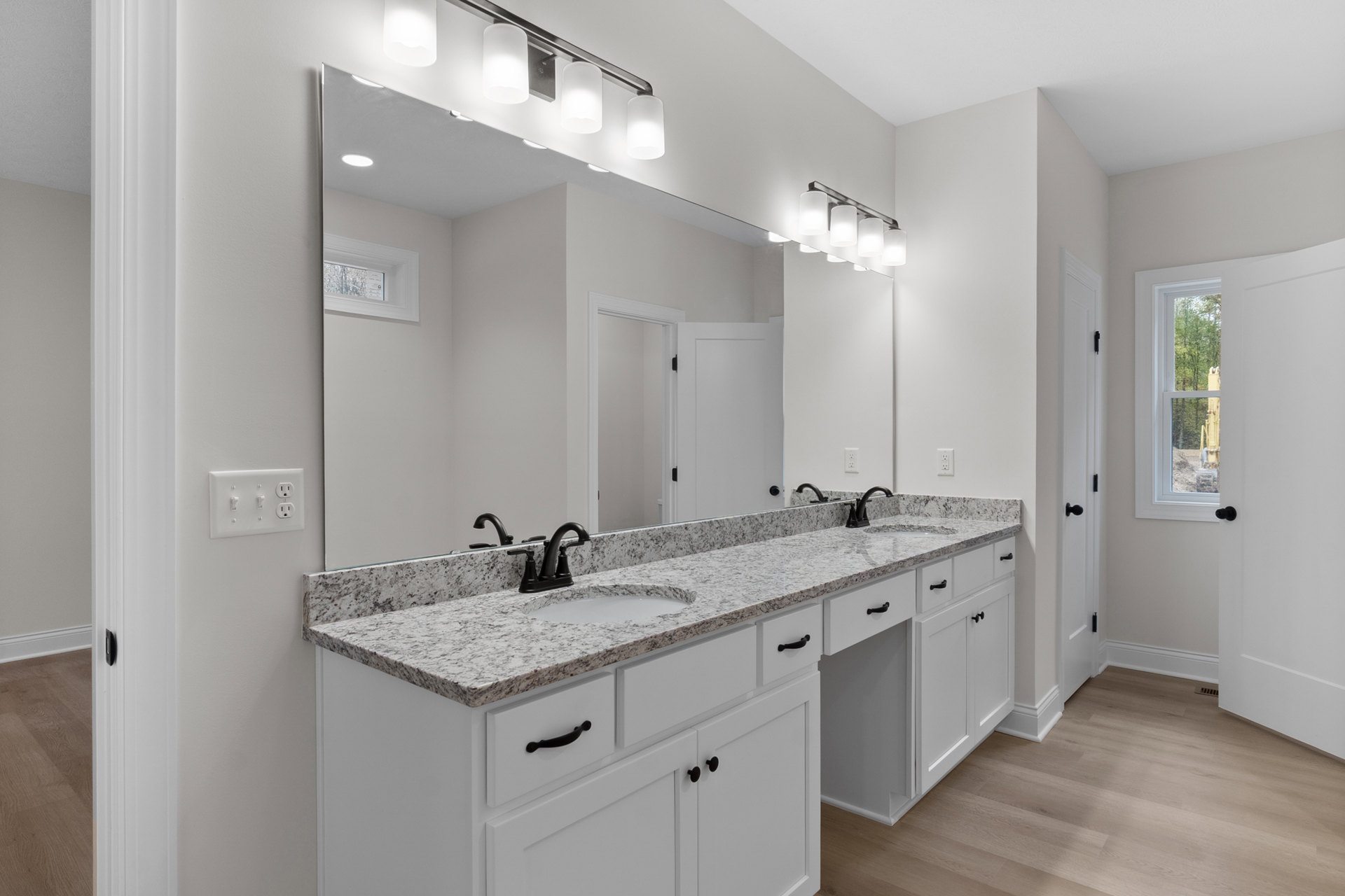 Bathroom with white tile walls, expansive mirror above a stone countertop, chrome faucet and sink, window overlooking green trees, modern light switch on wall, recessed ceiling