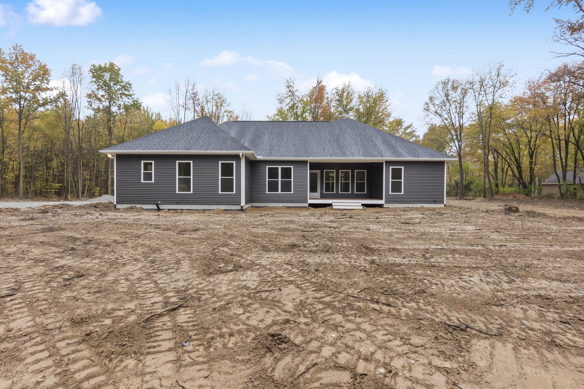 Single-story house with white-framed windows, covered porch, and unfinished dirt yard with tire tracks; trees and cloudy sky in background