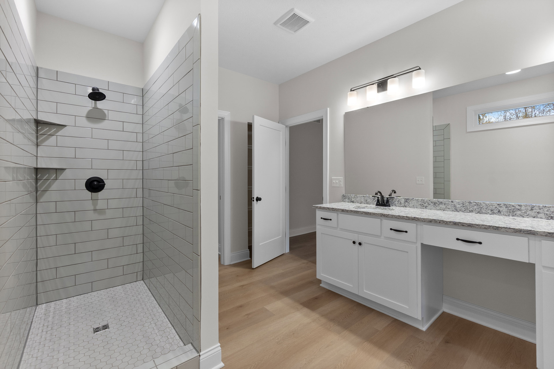 White tile shower with black shower head and handle, glass window showing clear sky, white door with black knob, light-colored floor, modern sink and countertop.