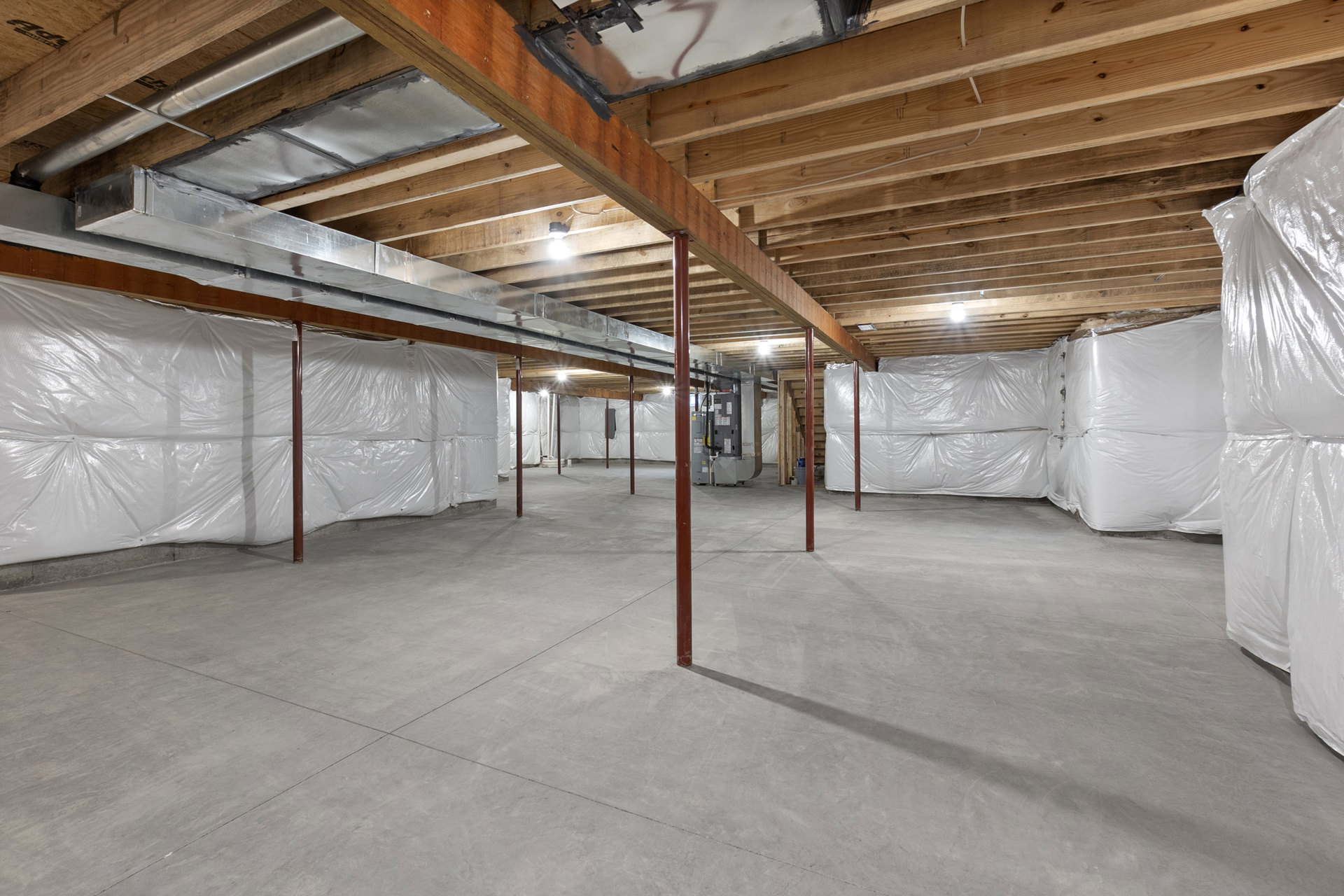 Basement room with white plastic coverings on walls and ceiling, exposed beams, concrete floor, and red support poles