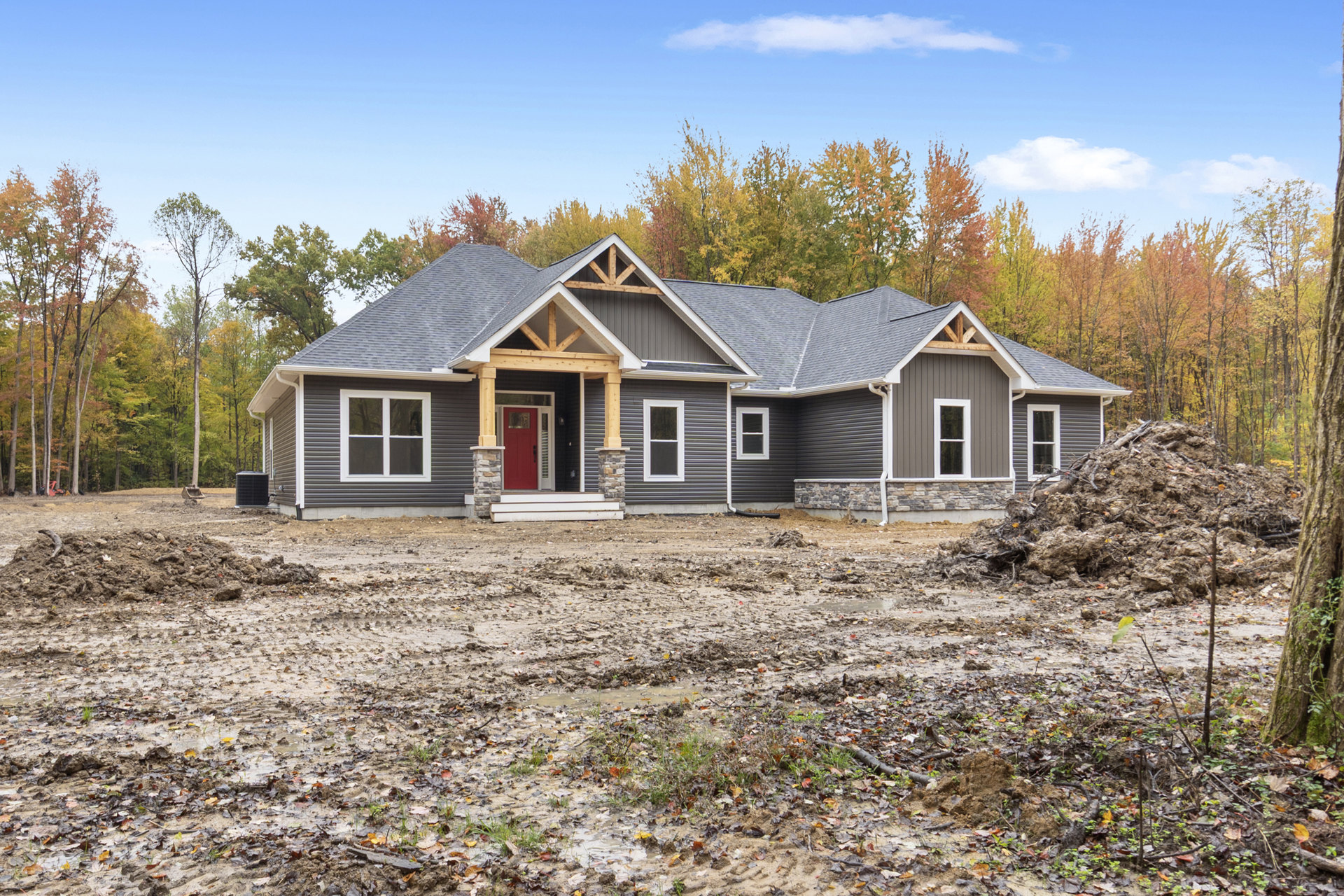 Partially built house with white-framed windows, red door with glass insert, exposed dirt yard, and trees with yellow leaves in the background
