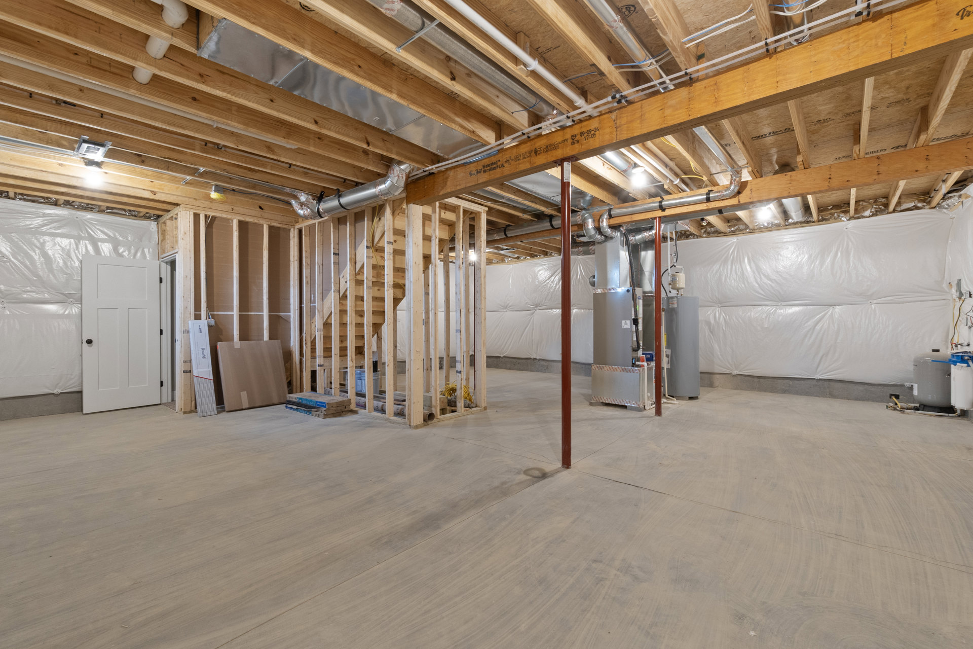 Basement room with exposed wood ceiling beams, visible pipes, concrete floor, red support pole, white door with black handle, cardboard box leaning against wall, staircase in