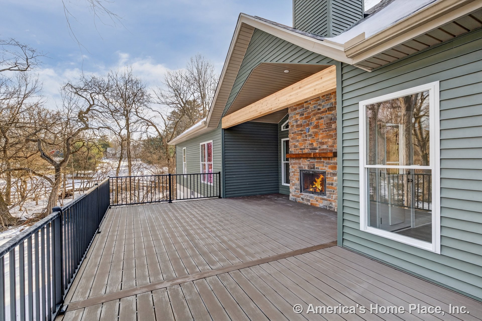 Covered composite deck with stone veneer fireplace, horizontal vinyl siding, black metal railing, large exterior windows, and wood porch ceiling.