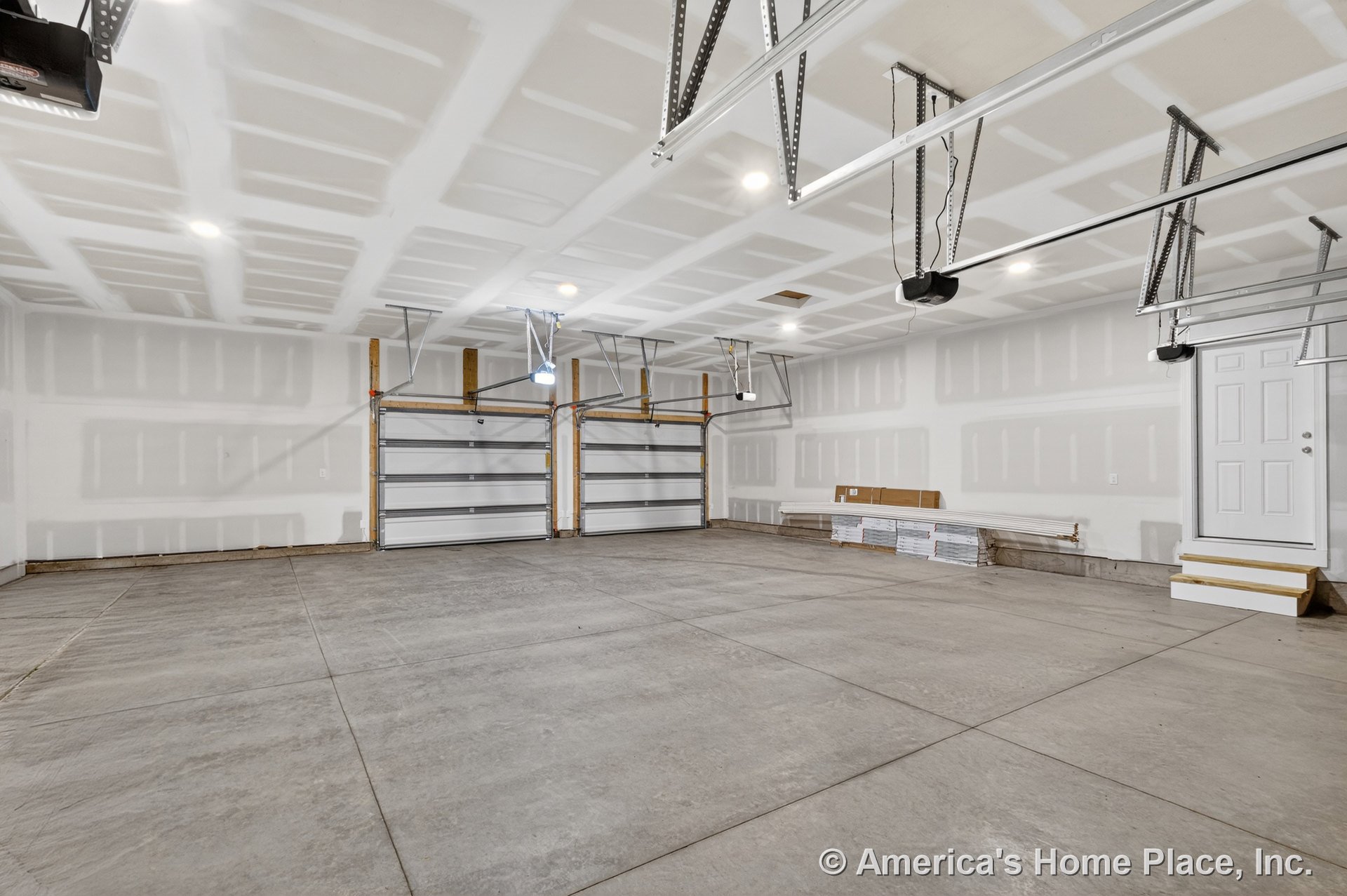 Unfinished drywall ceiling with built-in lighting, double overhead garage doors equipped with openers, concrete slab floor, side entry door accessed by steps, rear view of spacious