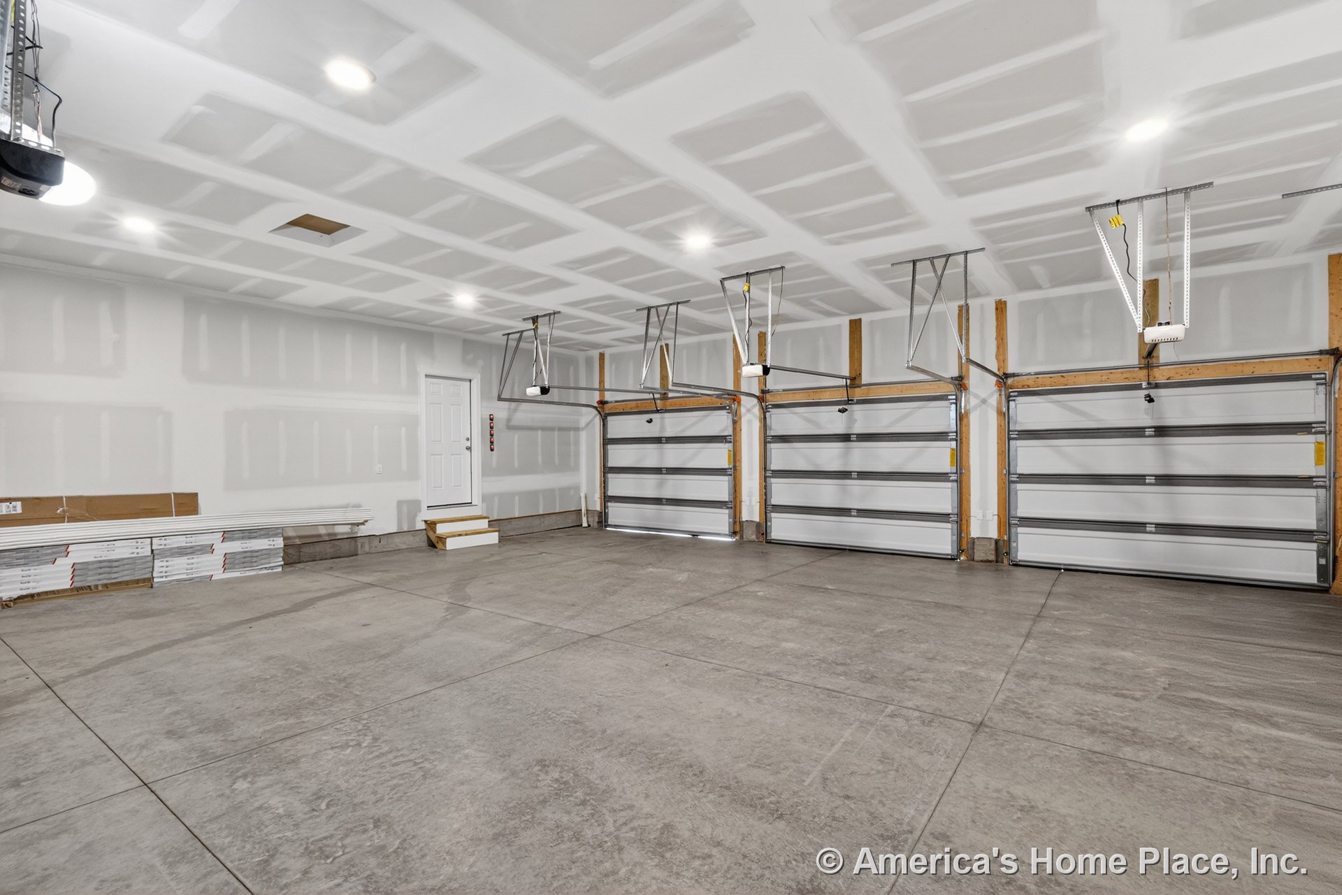 Three overhead garage doors with ceiling-mounted openers, unfinished drywall walls, concrete slab flooring, recessed ceiling lights, and an entry door with steps.
