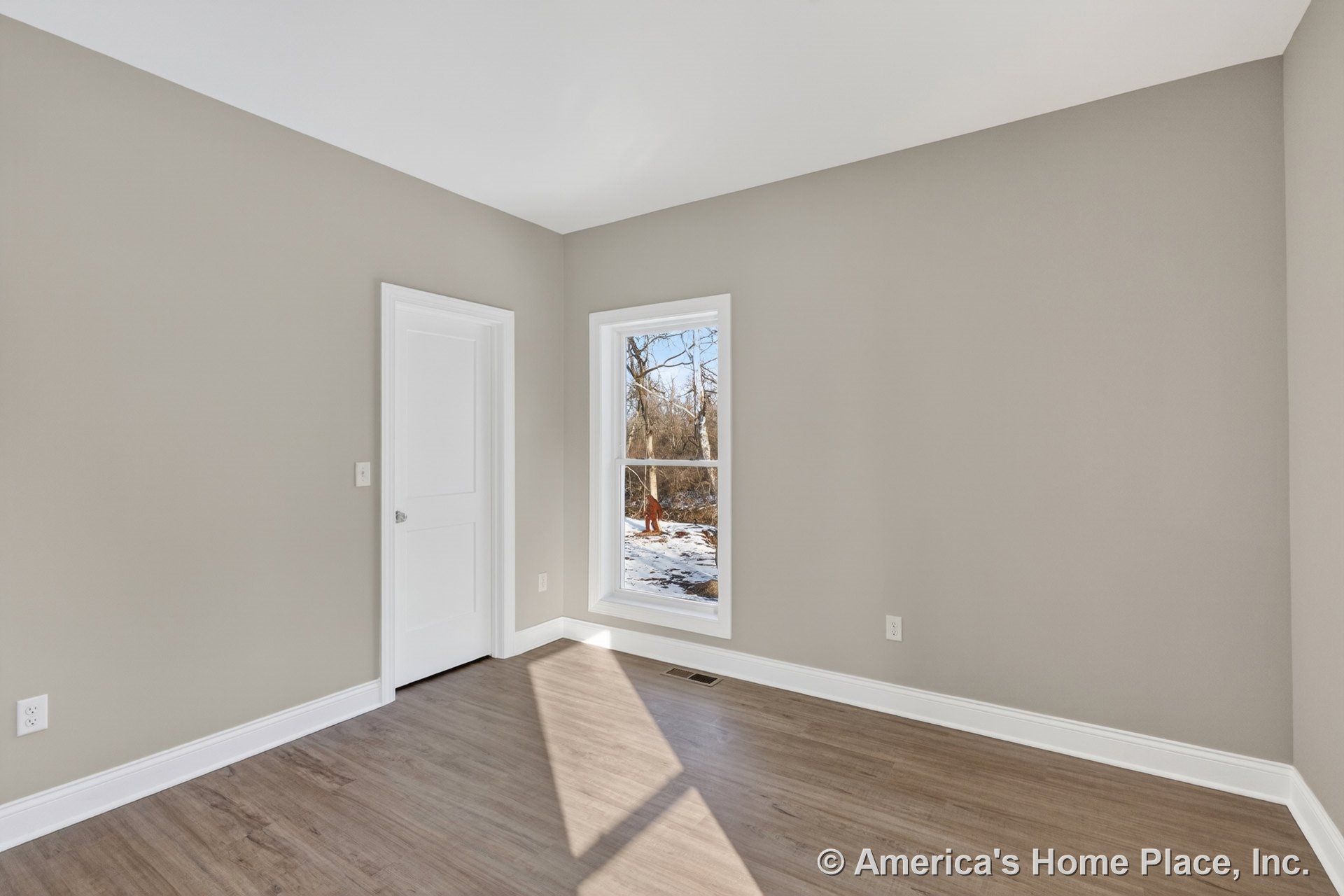 Bedroom with light wood plank flooring, large single window framed in white trim, neutral beige walls, white baseboards and door trim, white interior door, and flat white ceiling.