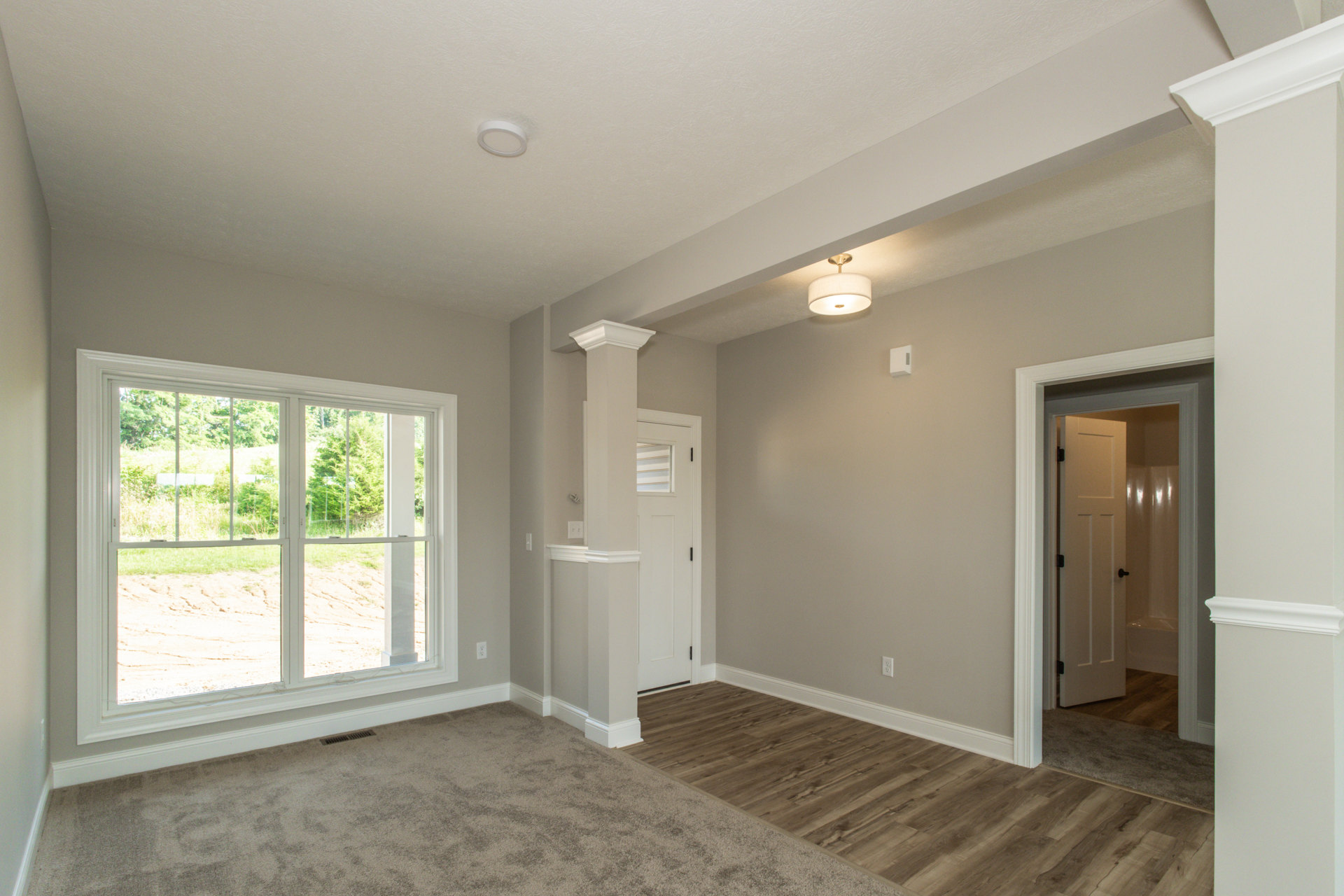 Wood floor with white walls and trim, ceiling-mounted white light fixture, window overlooking outdoors, door leading to hallway, doorway opening to bathroom with shower and bathtub