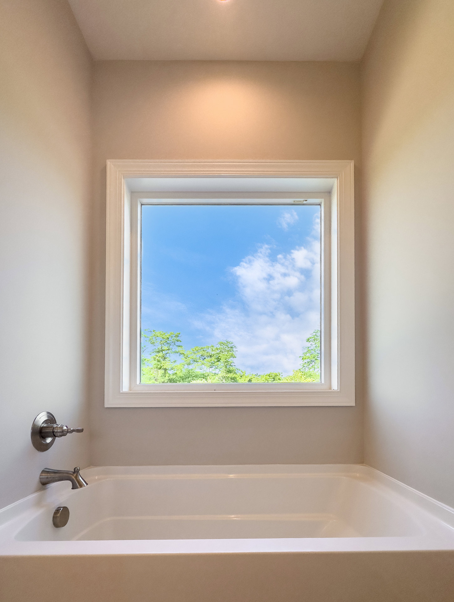 Freestanding white bathtub beneath large window with blue sky and tree view, brown and white walls, chrome faucet