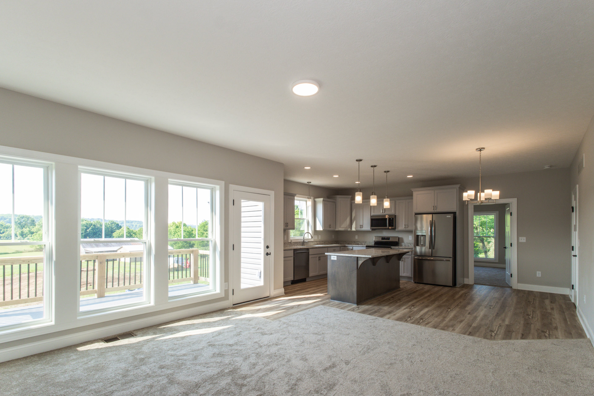 Open-concept kitchen and living room featuring a marble kitchen island, stainless steel refrigerator, ceiling light fixture, large windows, glass door to deck, and hardwood
