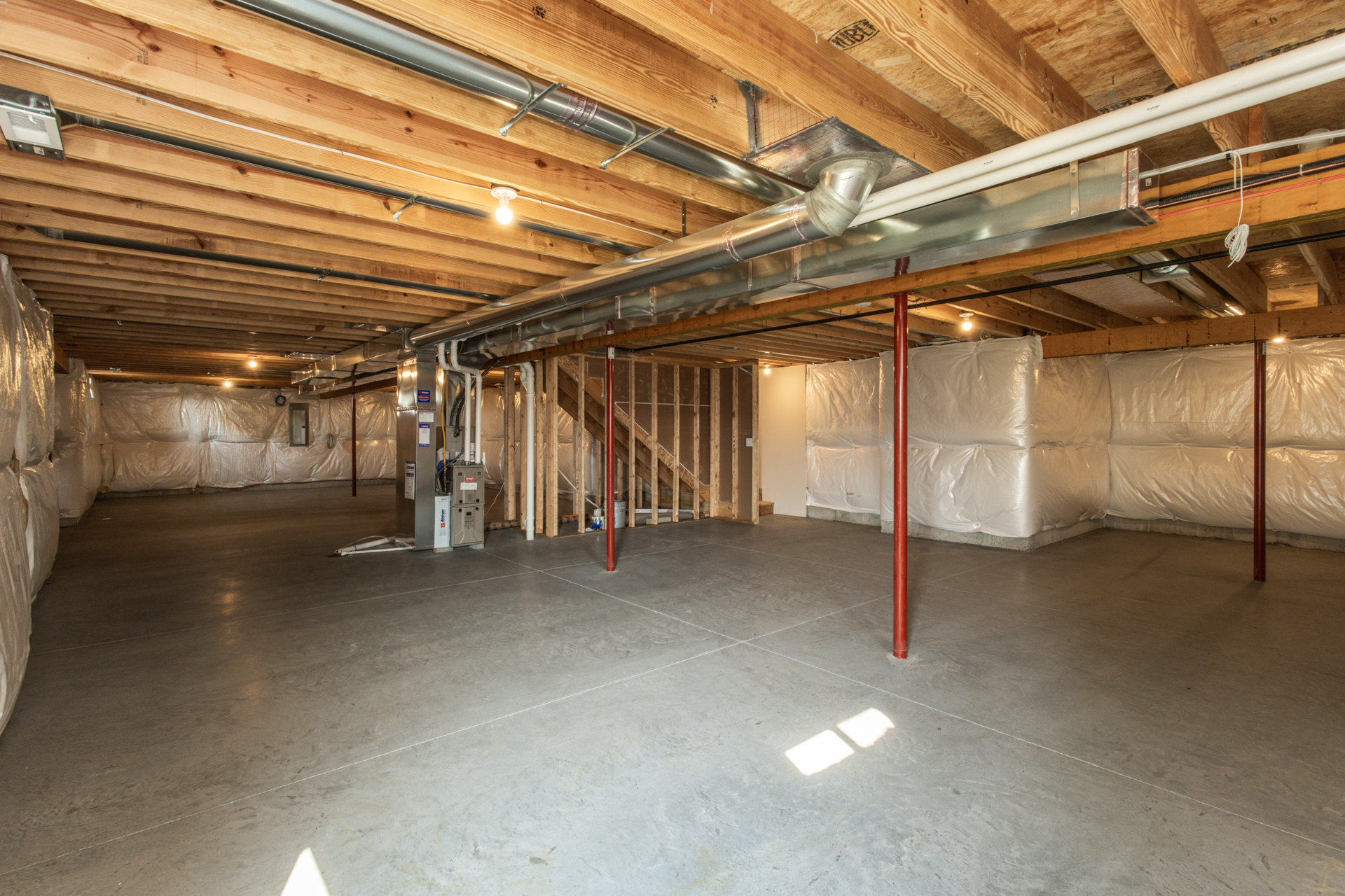 Basement room with exposed metal pipes along ceiling, concrete floor marked with white lines, large white insulation bags stacked near wall, overhead light illuminating white