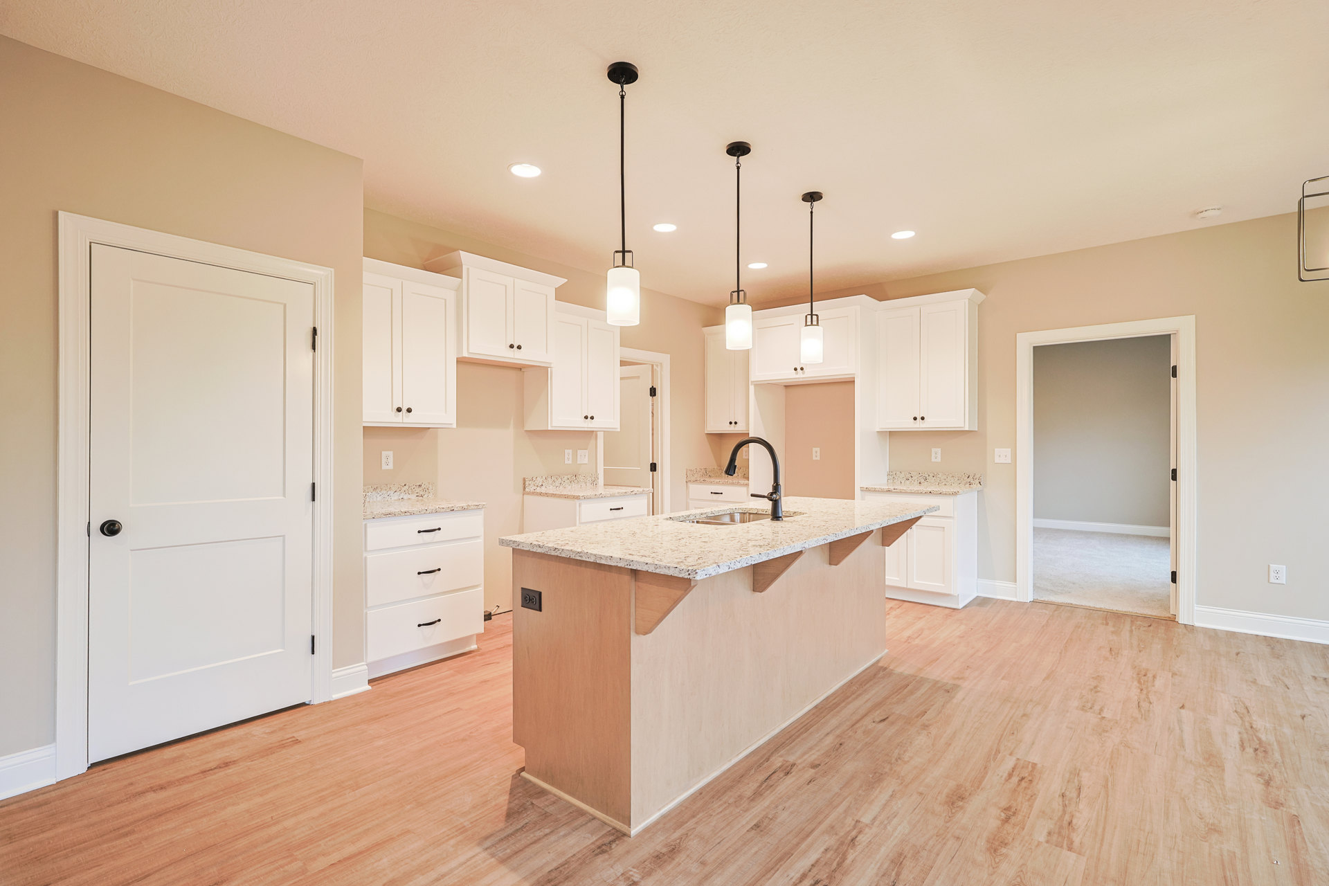 White kitchen with shaker cabinets, large central island featuring built-in sink, quartz countertops, tile flooring, recessed lighting, and a white door with black handle opening