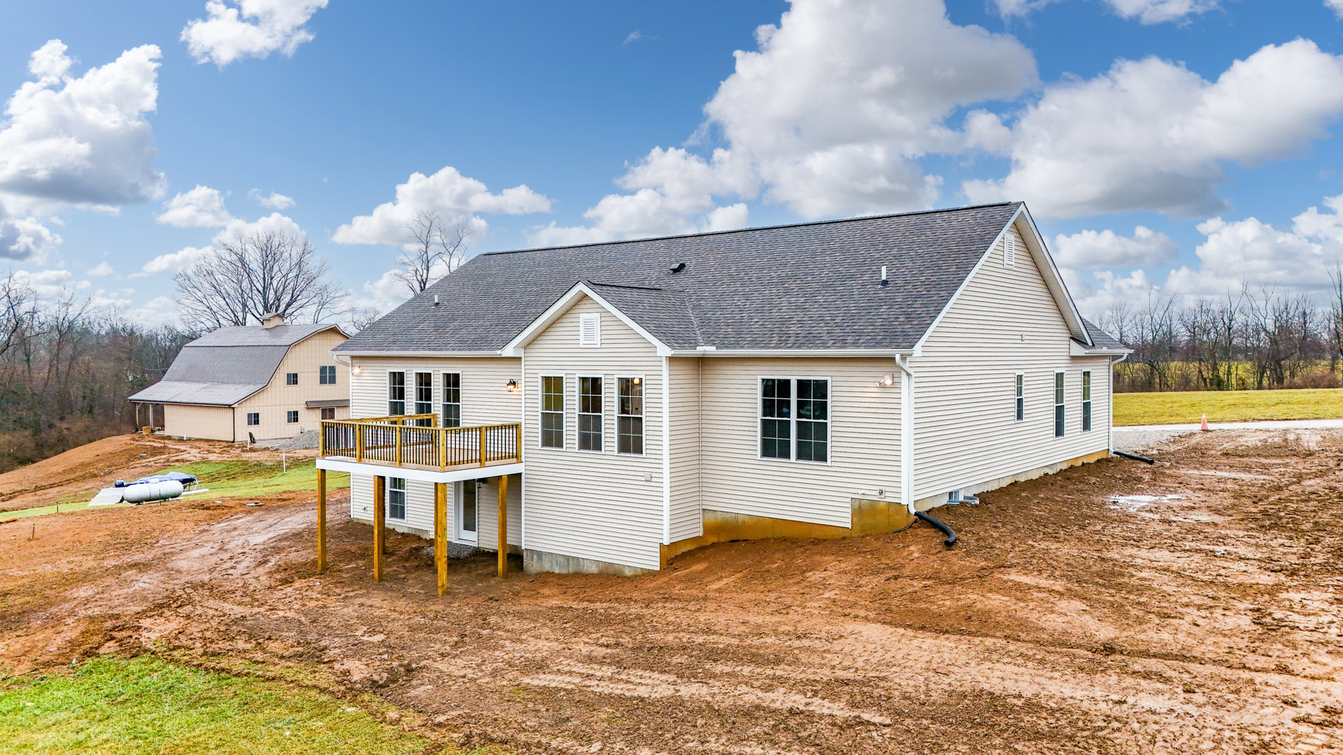 Wood deck with metal railing attached to a light-colored house, multi-pane windows, dirt yard in foreground, grassy hill and white barn with gray roof in background, blue sky with