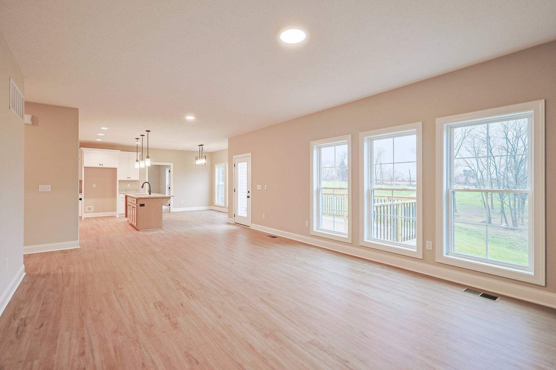 Spacious open-plan room featuring a wood floor, central kitchen island with sink, large windows overlooking trees, and recessed ceiling lighting.