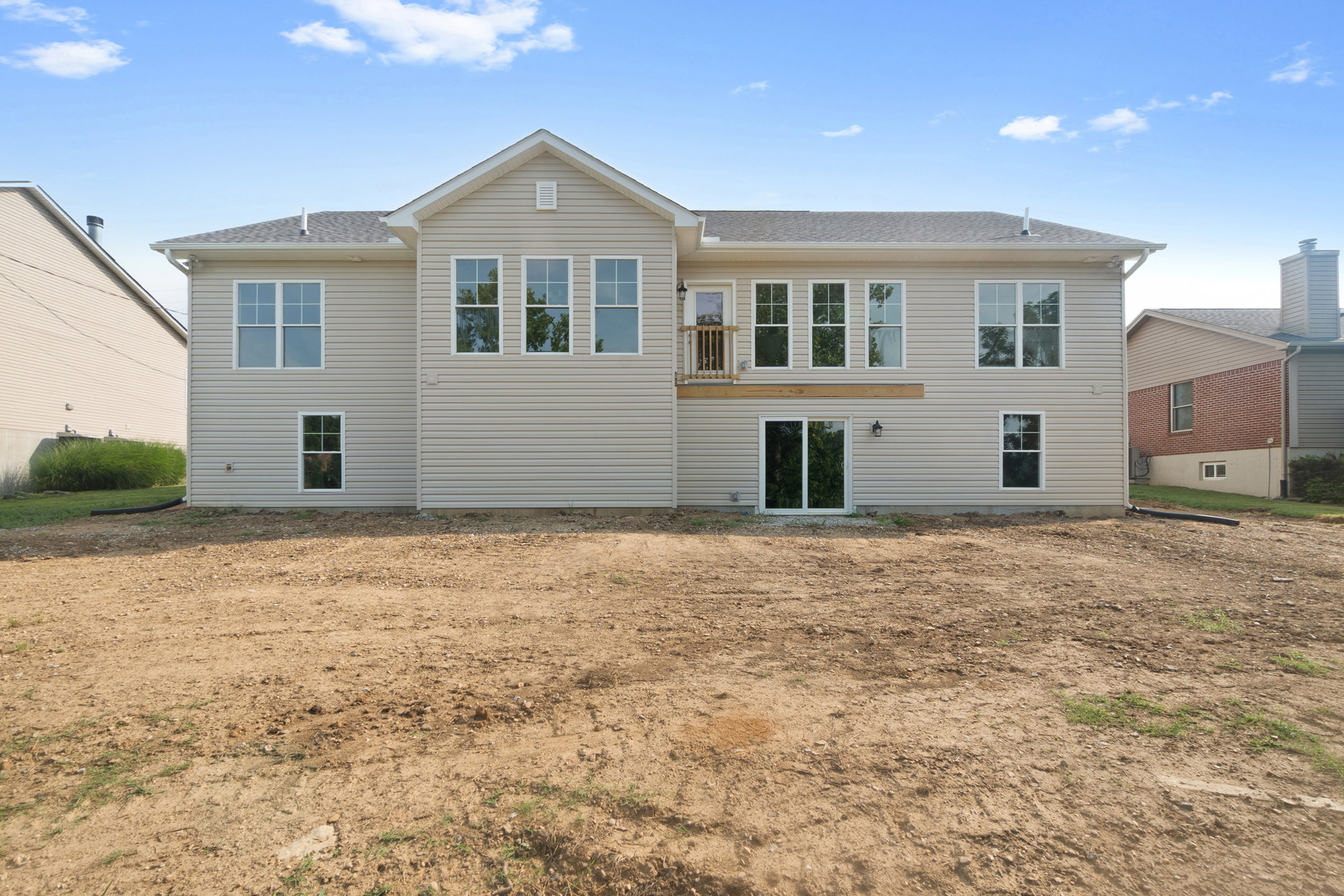 White modern house with three windows, balcony, white sliding glass door, and white siding, set beside a dirt field under a partly cloudy sky