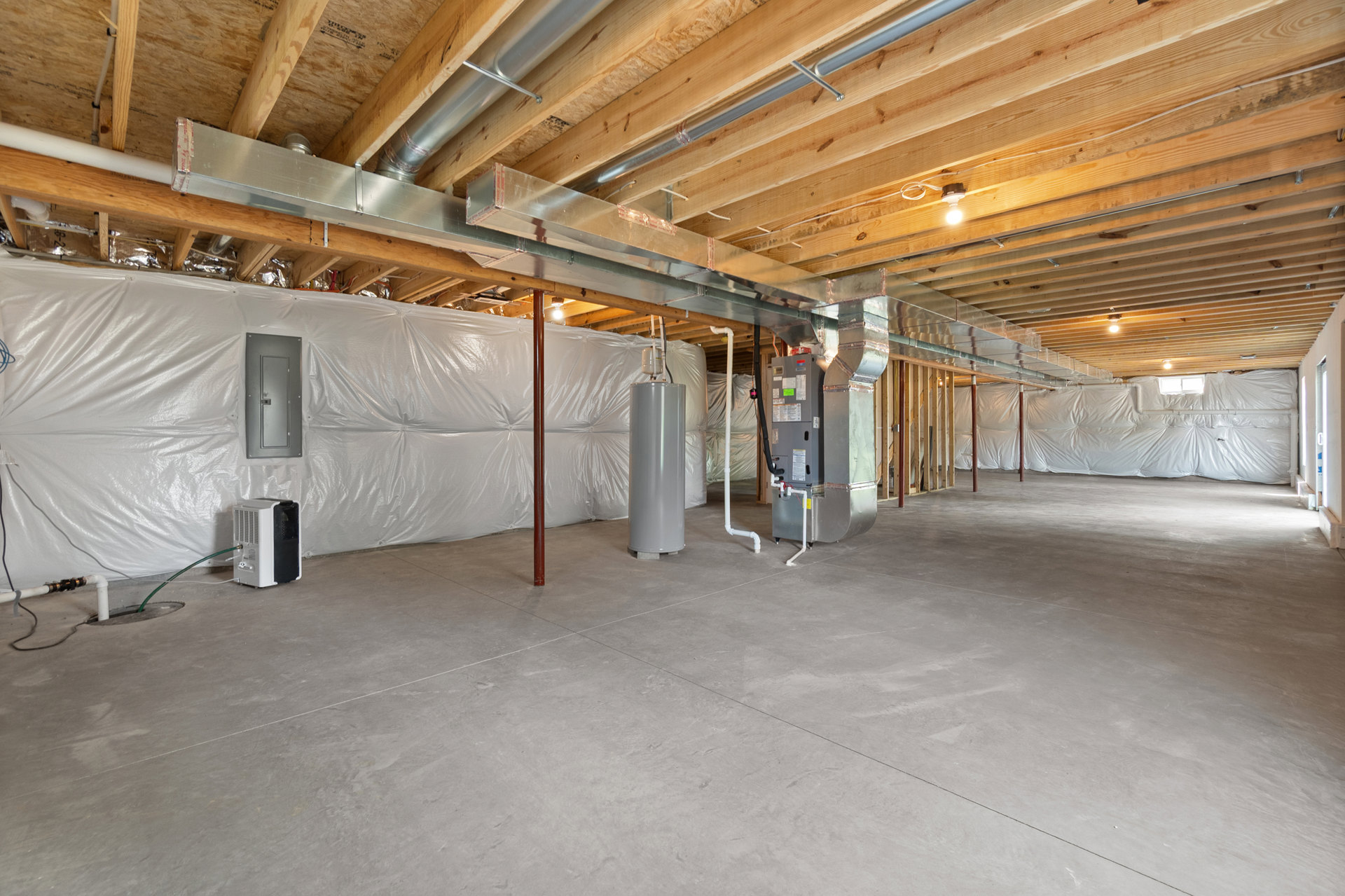 Basement room with exposed metal beams, visible pipes, grey metal door, concrete floor, white insulation covering walls, and heating equipment.
