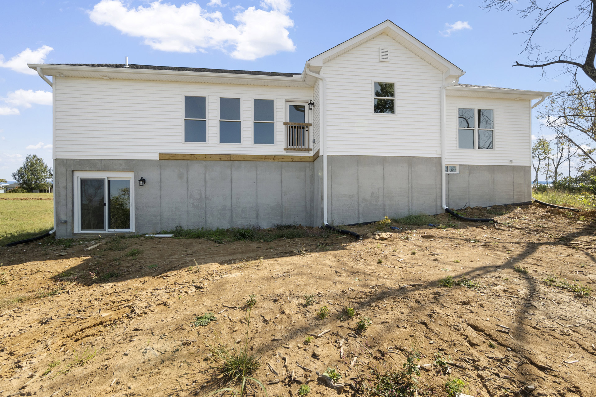 White house under construction with concrete foundation, white-framed windows reflecting trees, dirt hill and field with exposed white pipe, cloudy sky overhead.