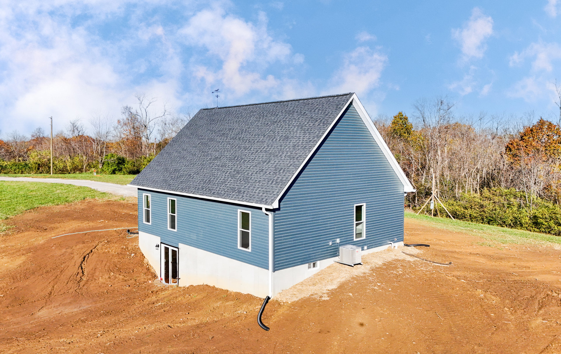 Partially built house with blue metal roof, exposed framing, dirt lot, portable white air conditioner, weather vane on roof, nearby trees, cloudy sky