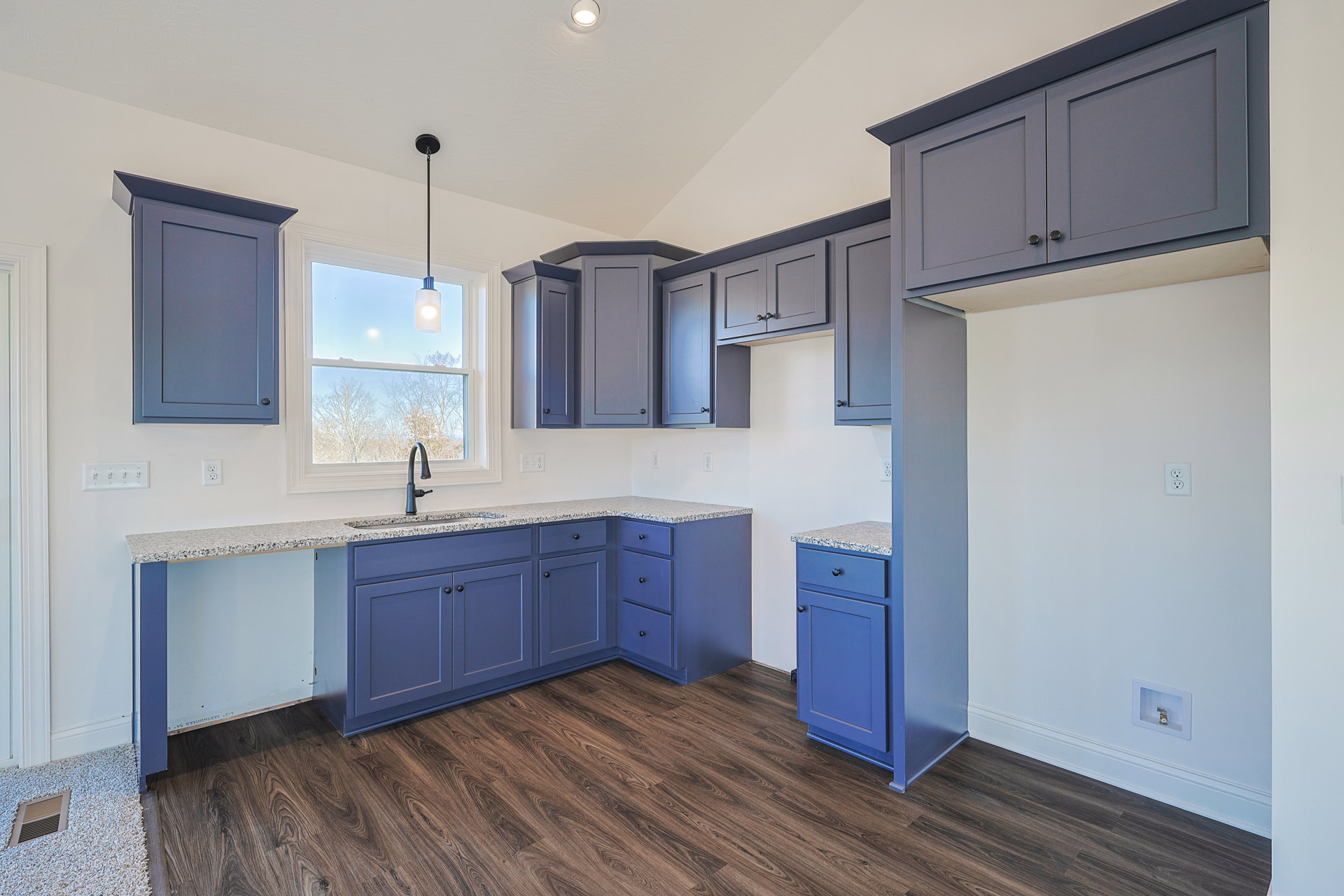Blue kitchen cabinets with black handles, wood flooring, black countertop, window above sink, and carpeted area with floor vent.