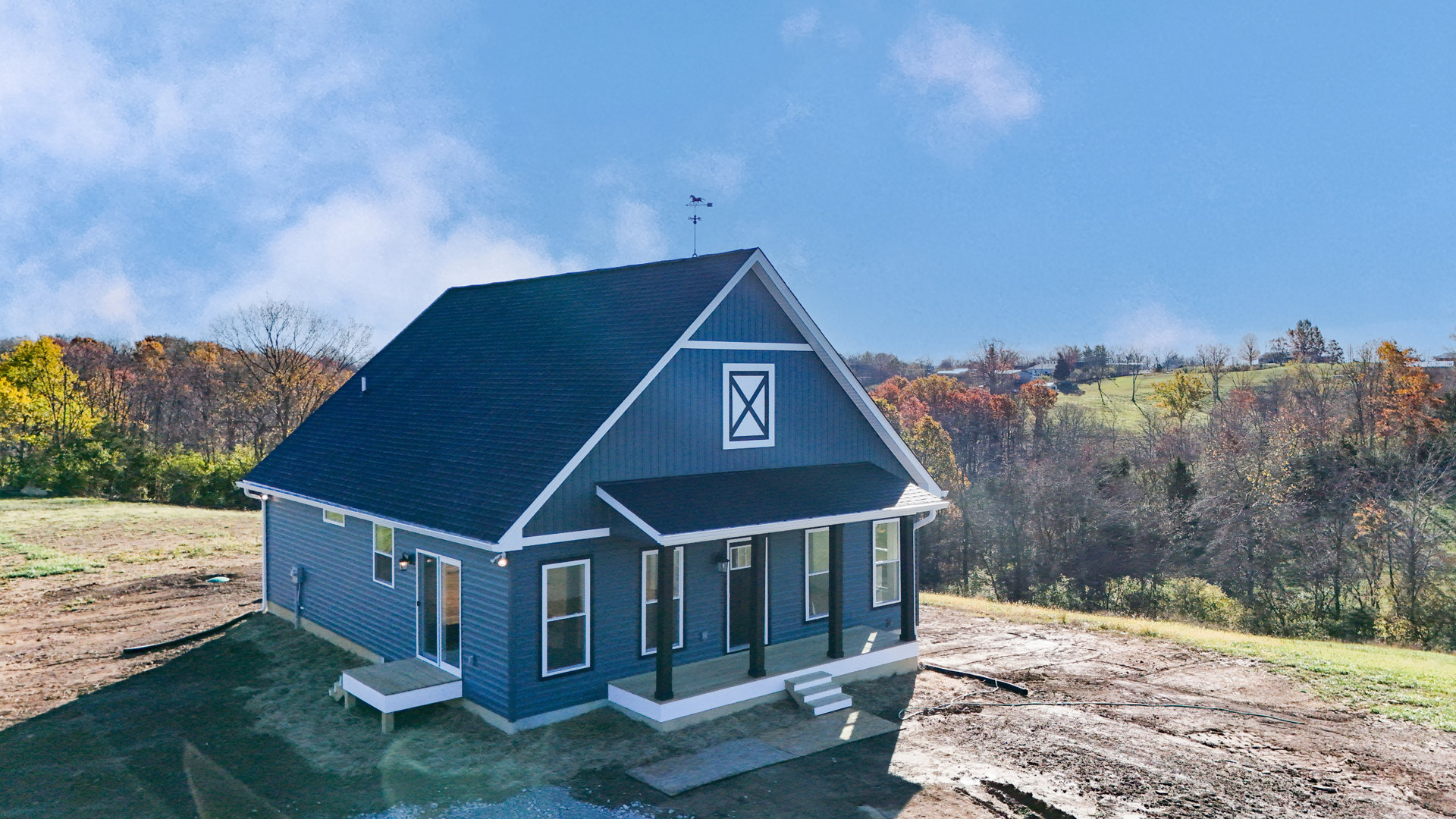 Blue metal roof atop a light blue cottage-style home, white-framed windows, weather vane on roof peak, blue and white sign near entry, surrounded by trees and cloudy sky.
