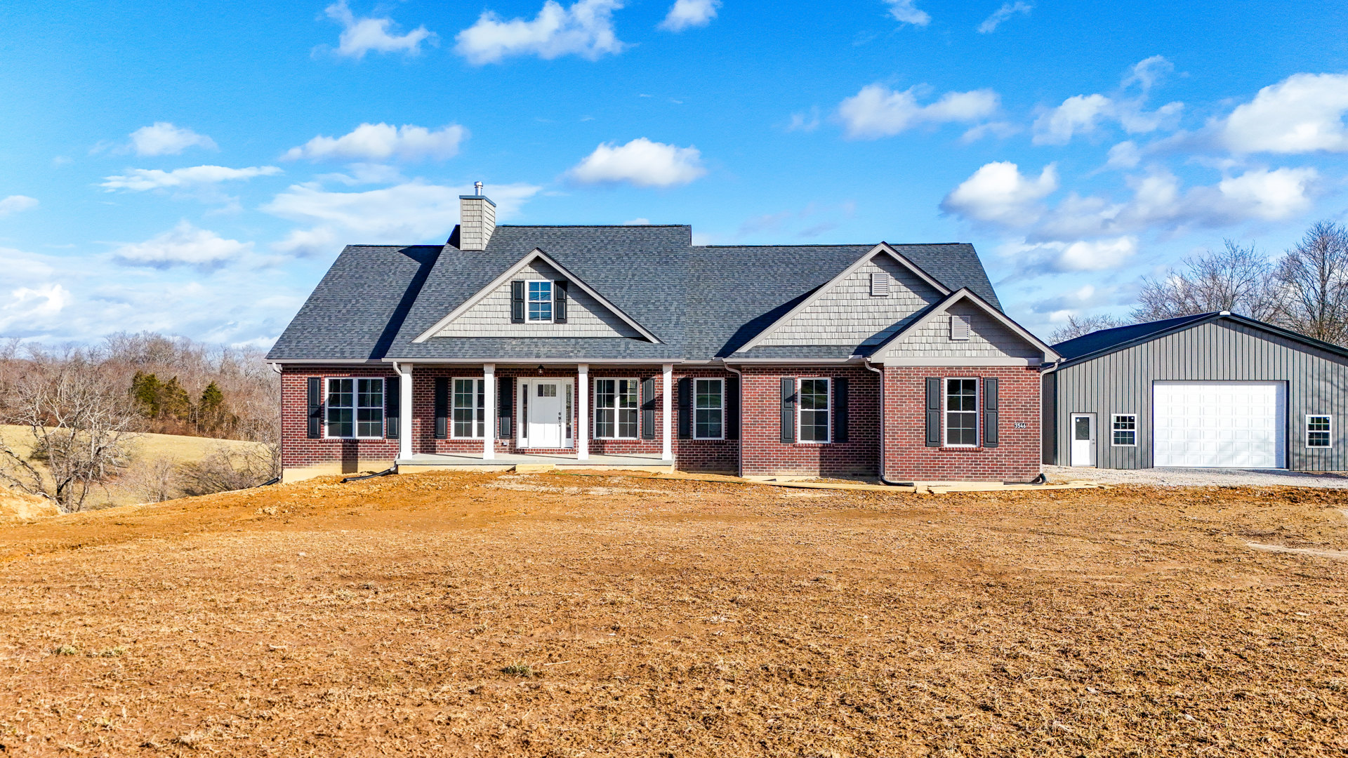 Brick house with white garage door and black roof, chimney, white entry door with window, large dirt field in foreground, cloudy sky above