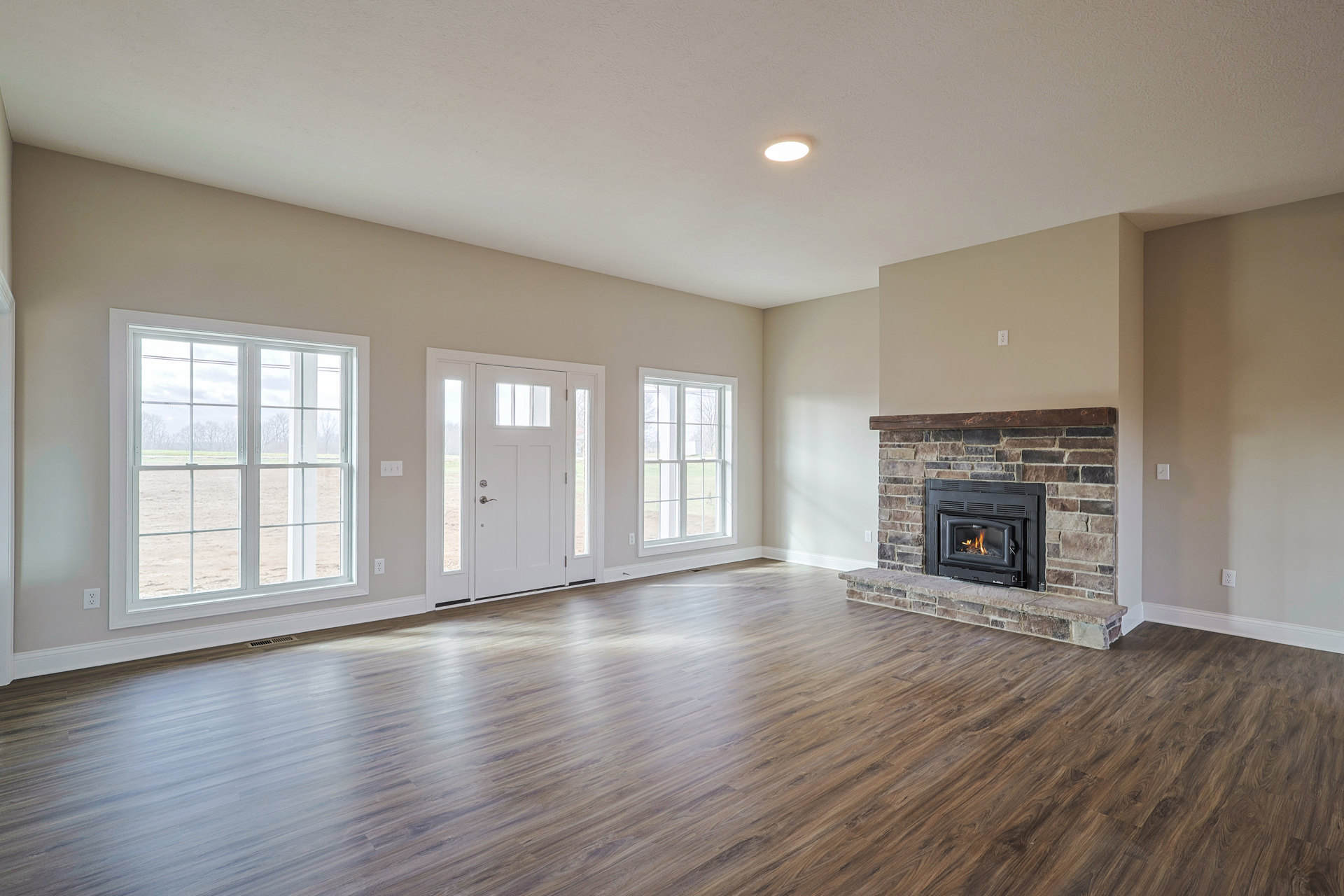 Living room with wood flooring, stone fireplace with burning fire and wood mantle, large window overlooking grassy field, white door with glass panes, neutral walls and ceiling.