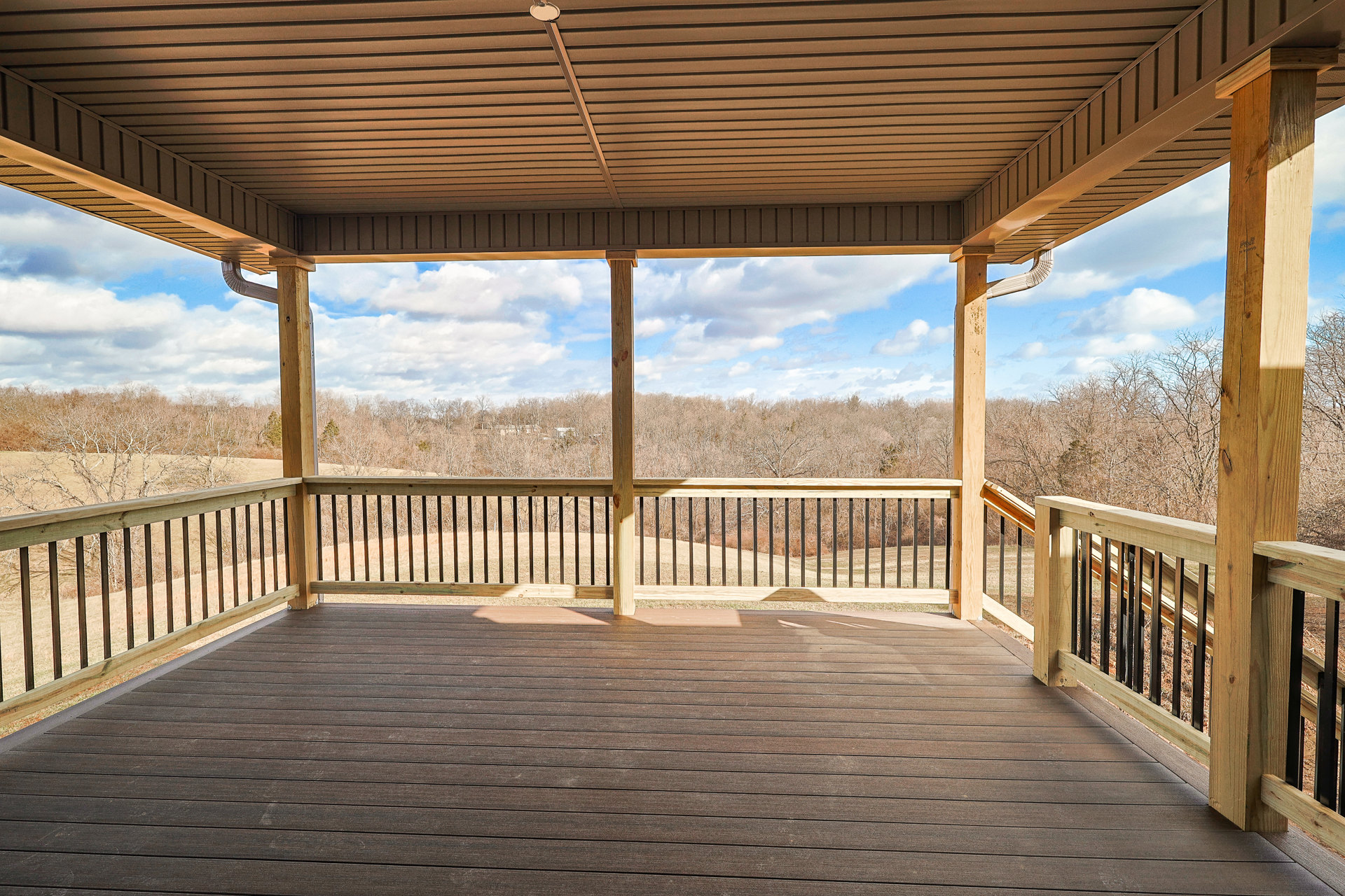 Wooden deck with horizontal railing overlooking leafy trees and bushes, partially shaded by ceiling, outdoor landscape visible beyond fence.