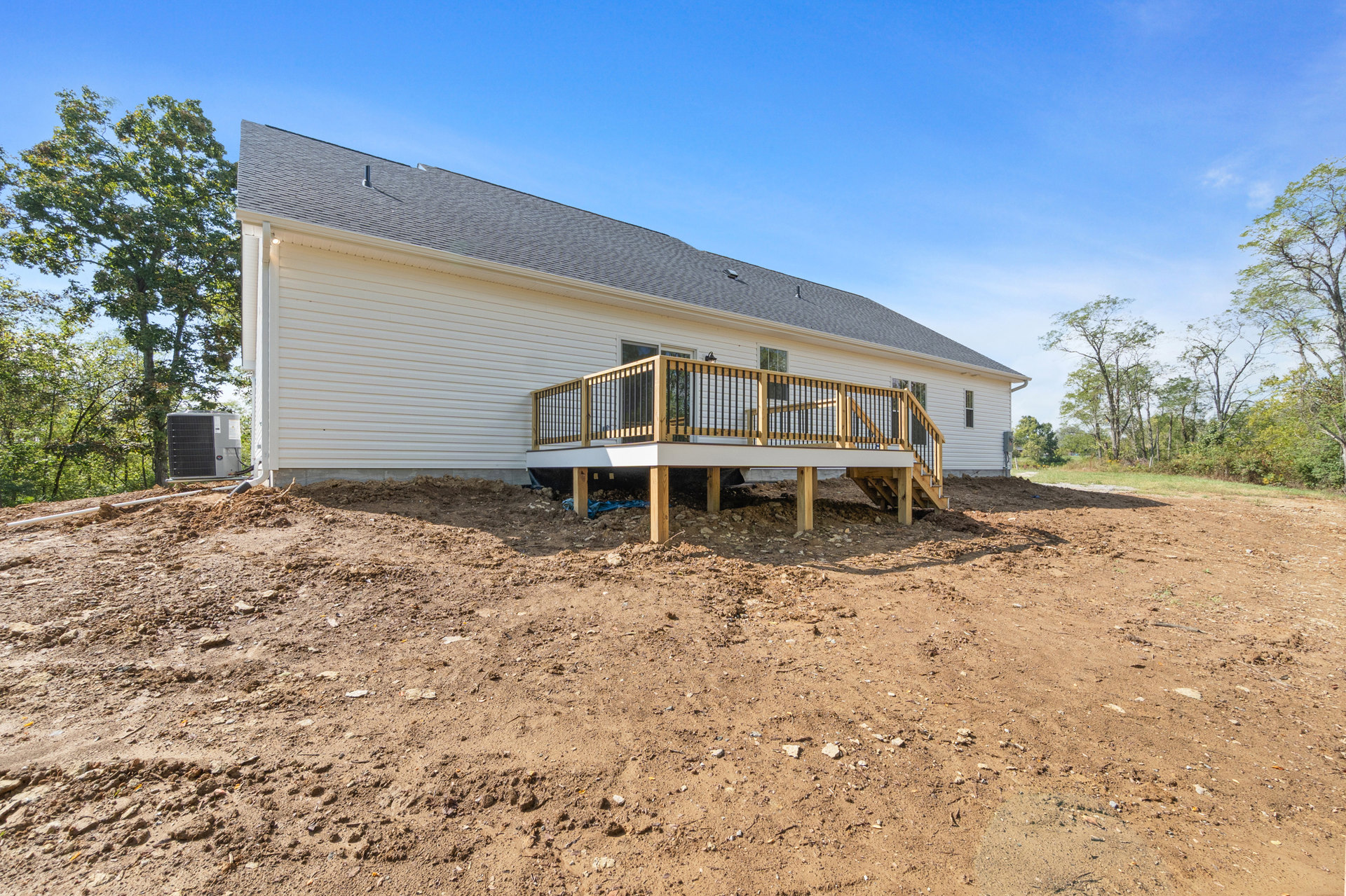 Wood deck with metal railing attached to a light-colored house, dirt and rocks covering the yard, leafy tree nearby, construction equipment visible
