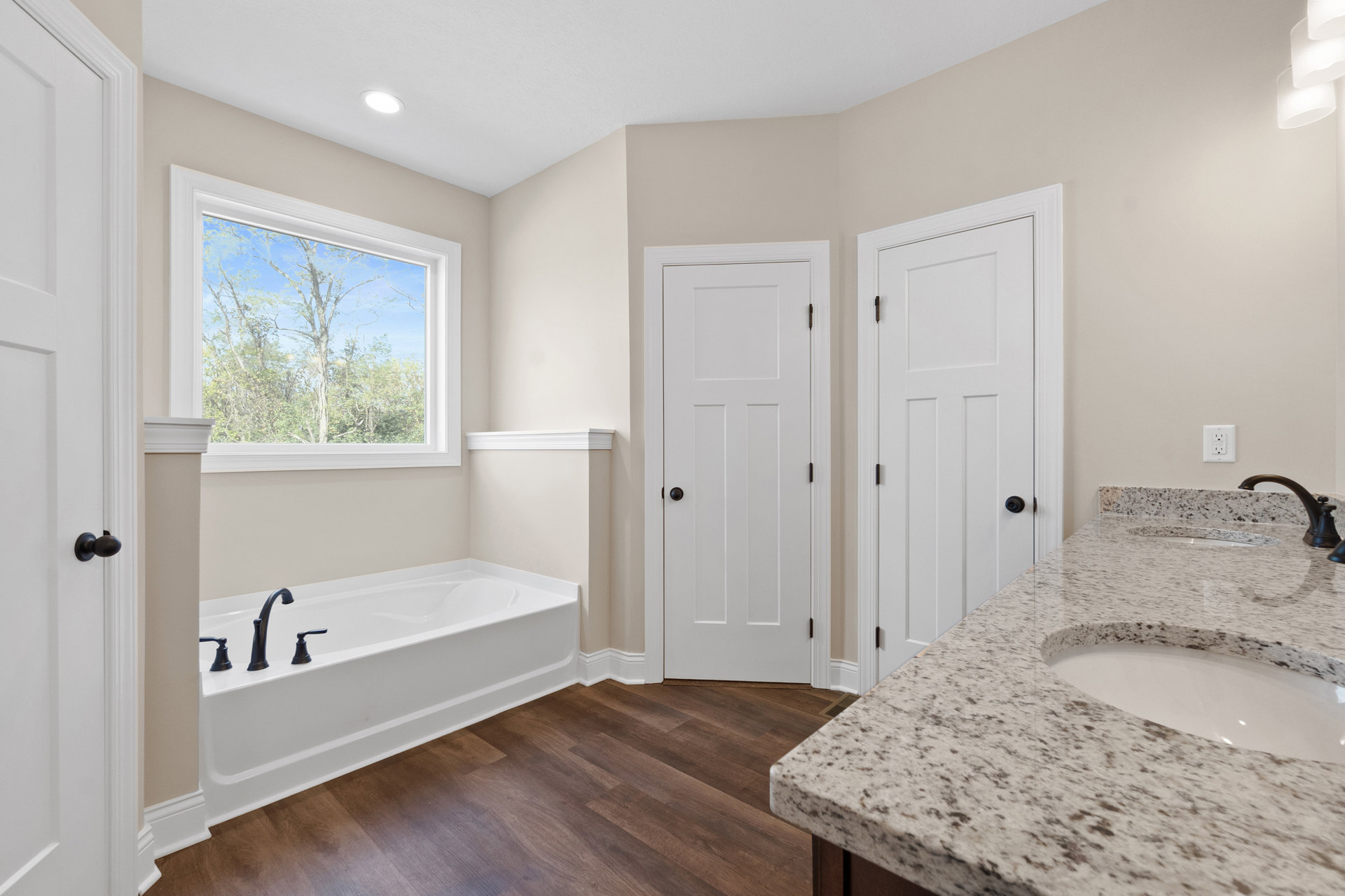 Bathroom with freestanding bathtub, wood floor, white countertop and sink, window overlooking trees, white door with black knob, chrome faucets