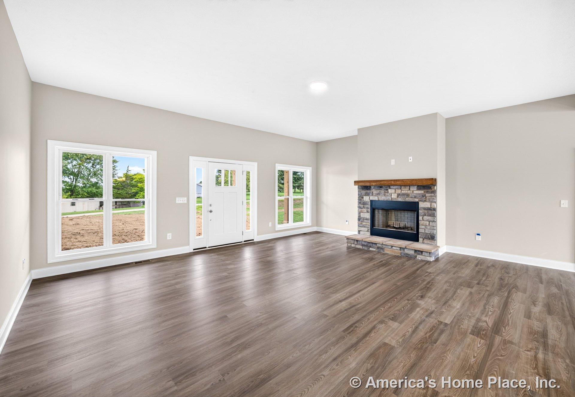 Stone fireplace with wood mantel centered on neutral painted wall, wide plank wood-look flooring, large double-hung windows with white trim and baseboards, recessed ceiling light