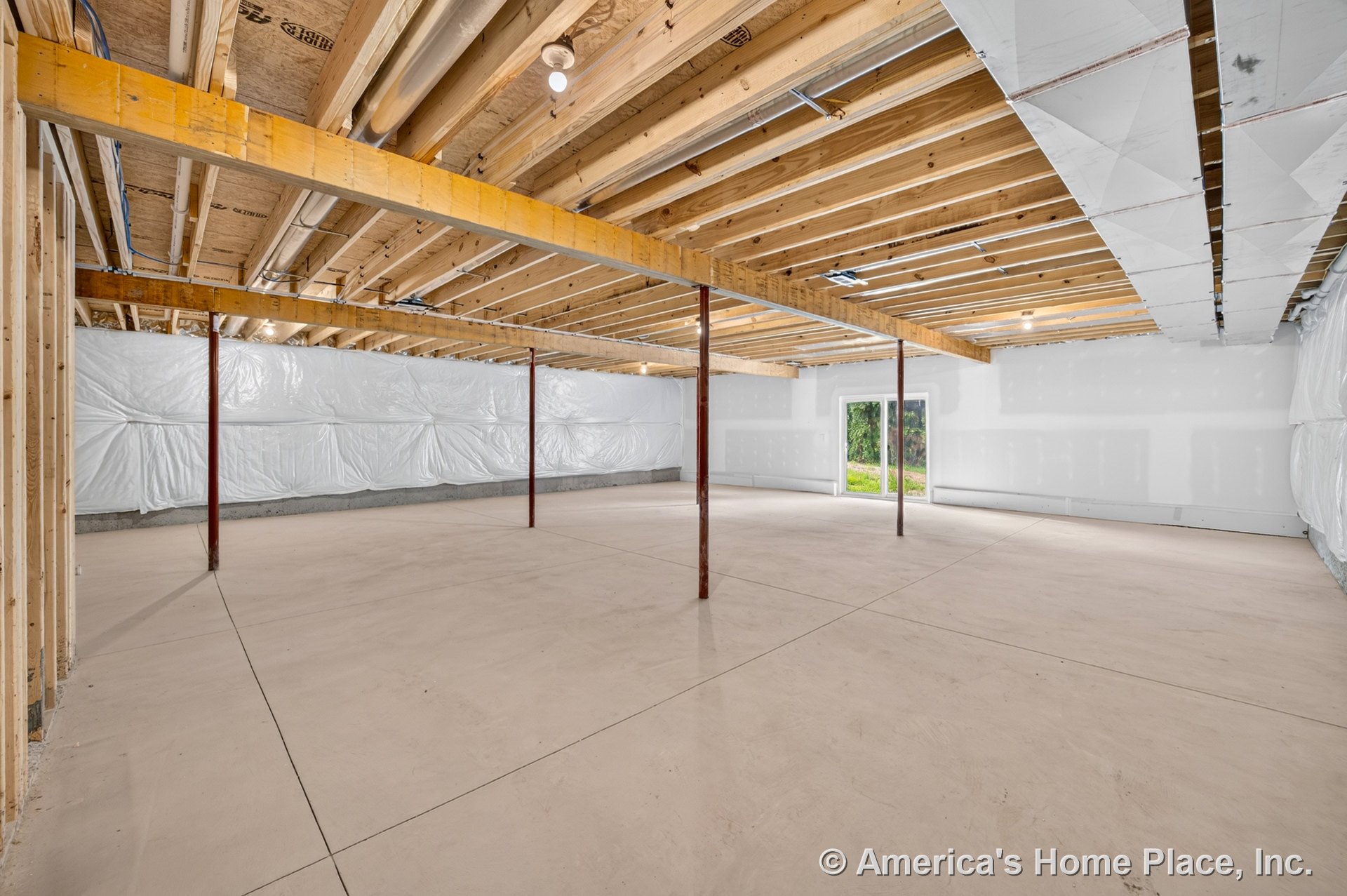 Unfinished basement with exposed wood ceiling joists, concrete slab floor, insulated wall panels, structural support columns, drywall partition, and walkout door allowing natural