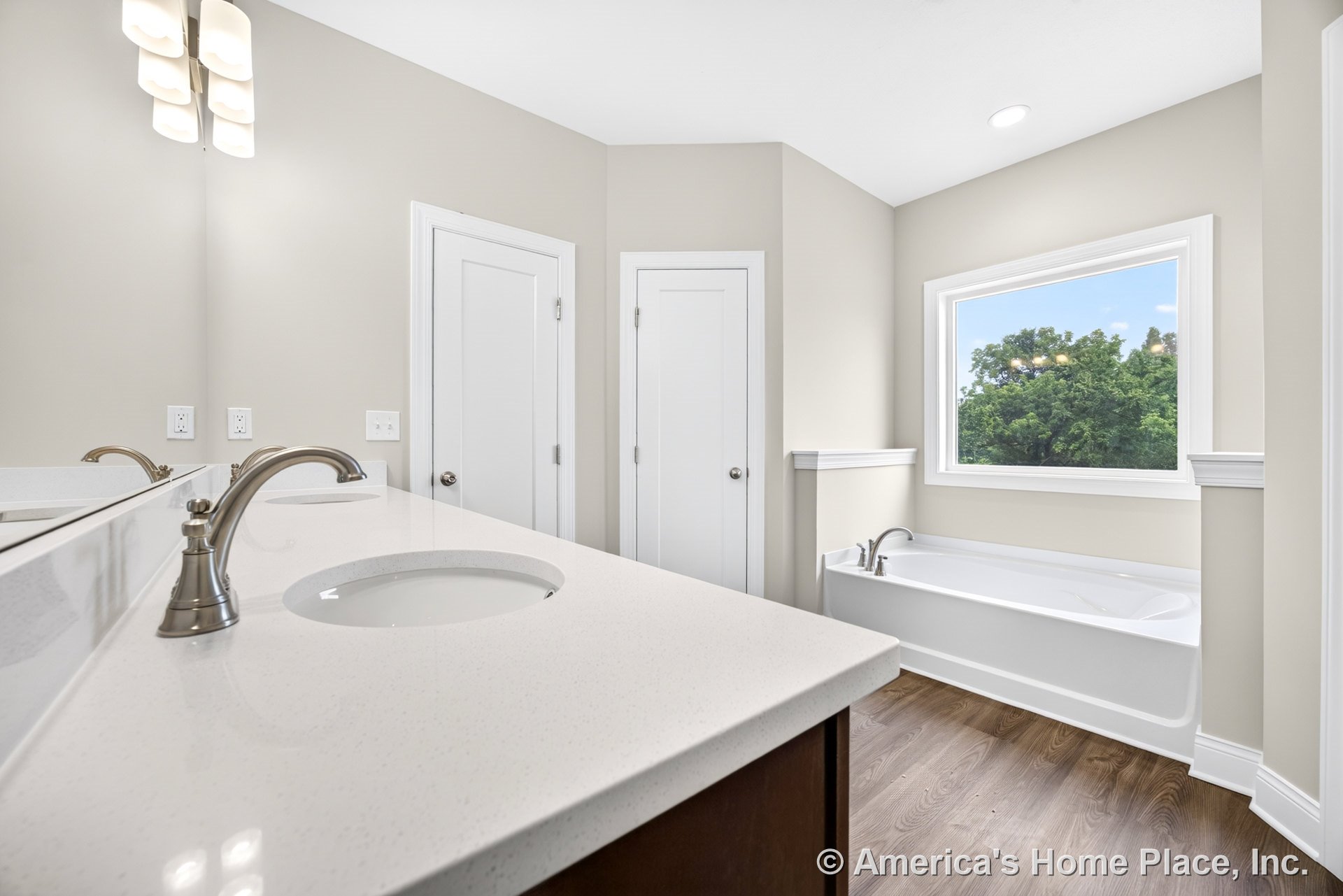 Built-in soaking tub positioned beneath a large window, double vanity with quartz countertops and brushed nickel faucets, wood-look flooring, white paneled doors, modern wall