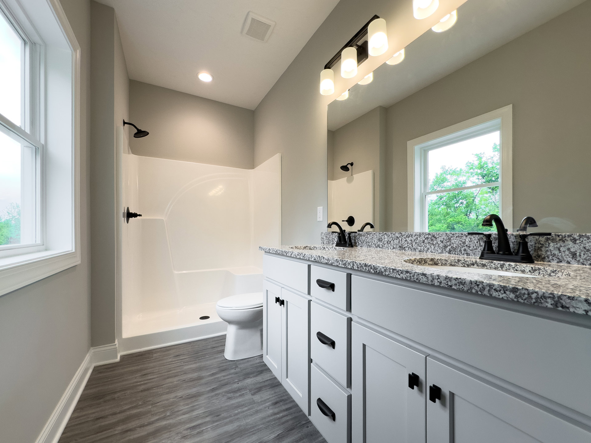 Bathroom with grey wood floor, white walls, marble countertop with black faucet, white sink, white toilet, glass shower enclosure, window overlooking trees.