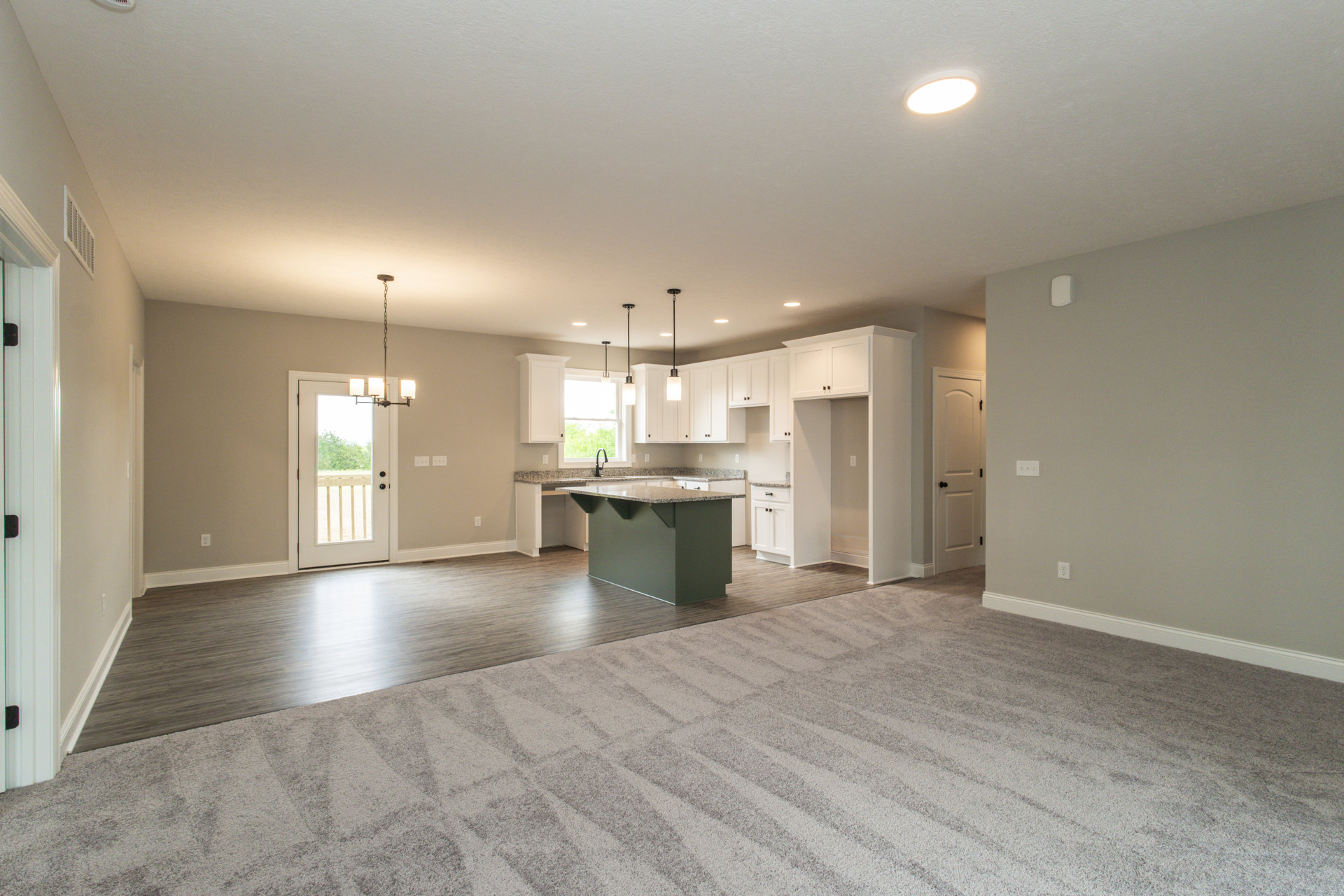 Open-concept kitchen and living room with carpeted flooring, granite kitchen island, ceiling light fixture, cabinetry, and a door leading to a deck with tree views.